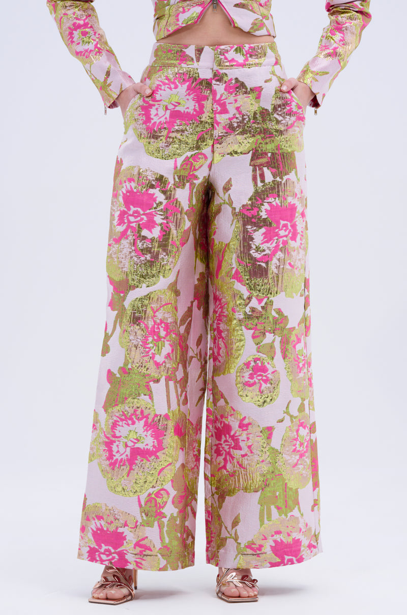 A person wearing the LOVELY FLORAL BROCADE TROUSER, featuring a wide leg and vibrant pink and green metallic floral details, with a matching top partially visible and metallic sandals, stands against a plain white background.