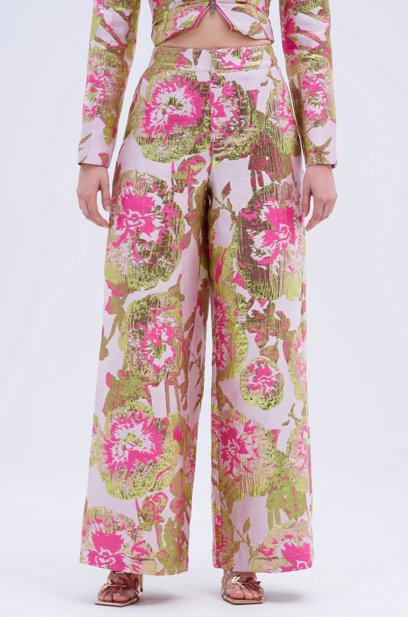 A person displays the wide-leg silhouette of LOVELY FLORAL BROCADE TROUSER, featuring bold pink and green florals with metallic detailing, paired with a matching top. Only the lower torso and legs are shown.