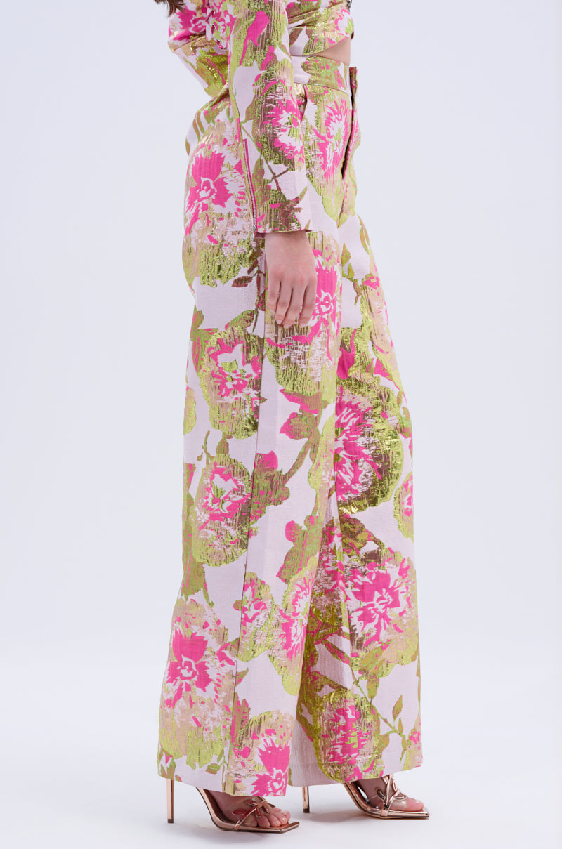 A person stands sideways, showing only the lower body in LOVELY FLORAL BROCADE TROUSER with a wide-leg silhouette and pink, green, and white patterns, paired with strappy high-heeled sandals.