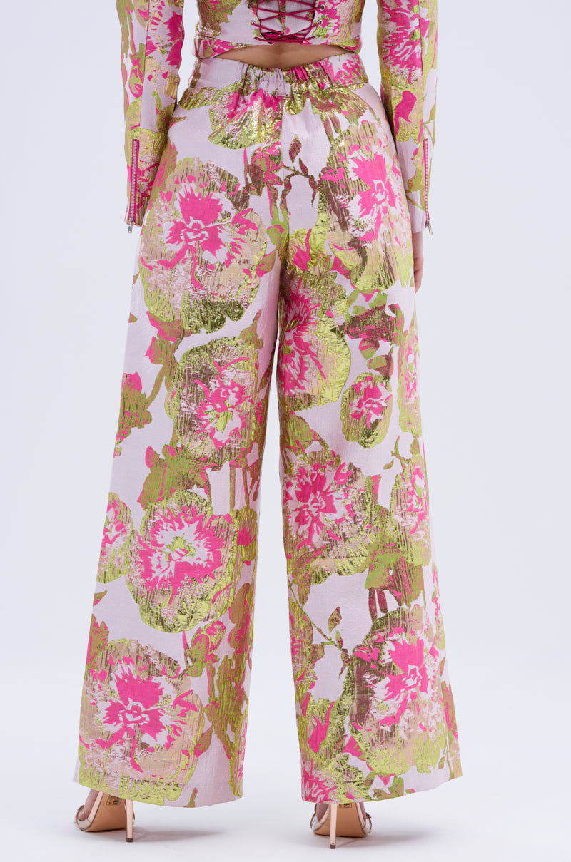 A person is shown from the back against a plain light background wearing LOVELY FLORAL BROCADE TROUSER, featuring a wide-leg silhouette and bold pink, green, and white florals, paired with open-toe heels.