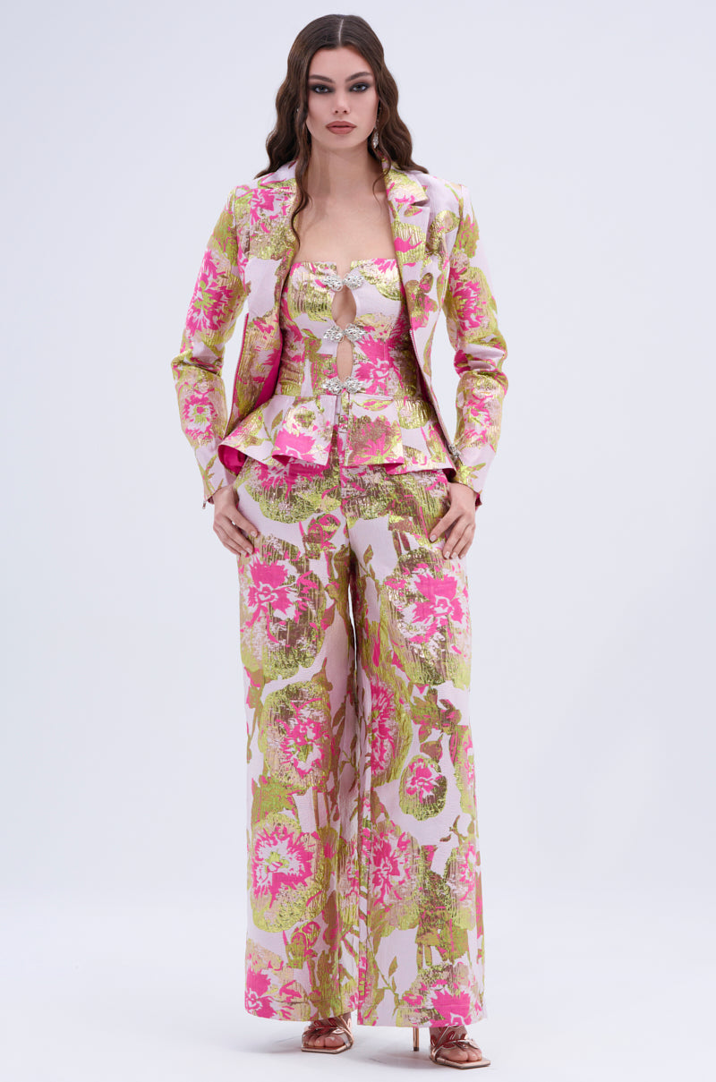 A woman stands facing forward in the LOVELY FLORAL BROCADE TROUSER set, a fitted blazer and peplum top paired with wide-leg trousers featuring pink, green, and gold metallic floral details. She has long wavy hair and wears open-toe heels.
