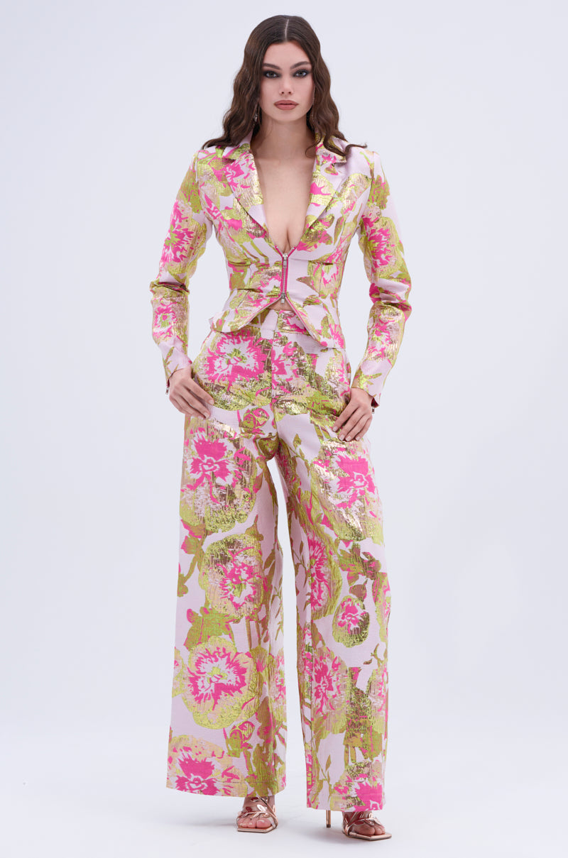 A woman stands confidently in the LOVELY FLORAL BROCADE TROUSER suit, showcasing a deep neckline and wide-leg silhouette with pink, green, and gold metallic floral accents. She completes the look with metallic pink heels against a plain light background.