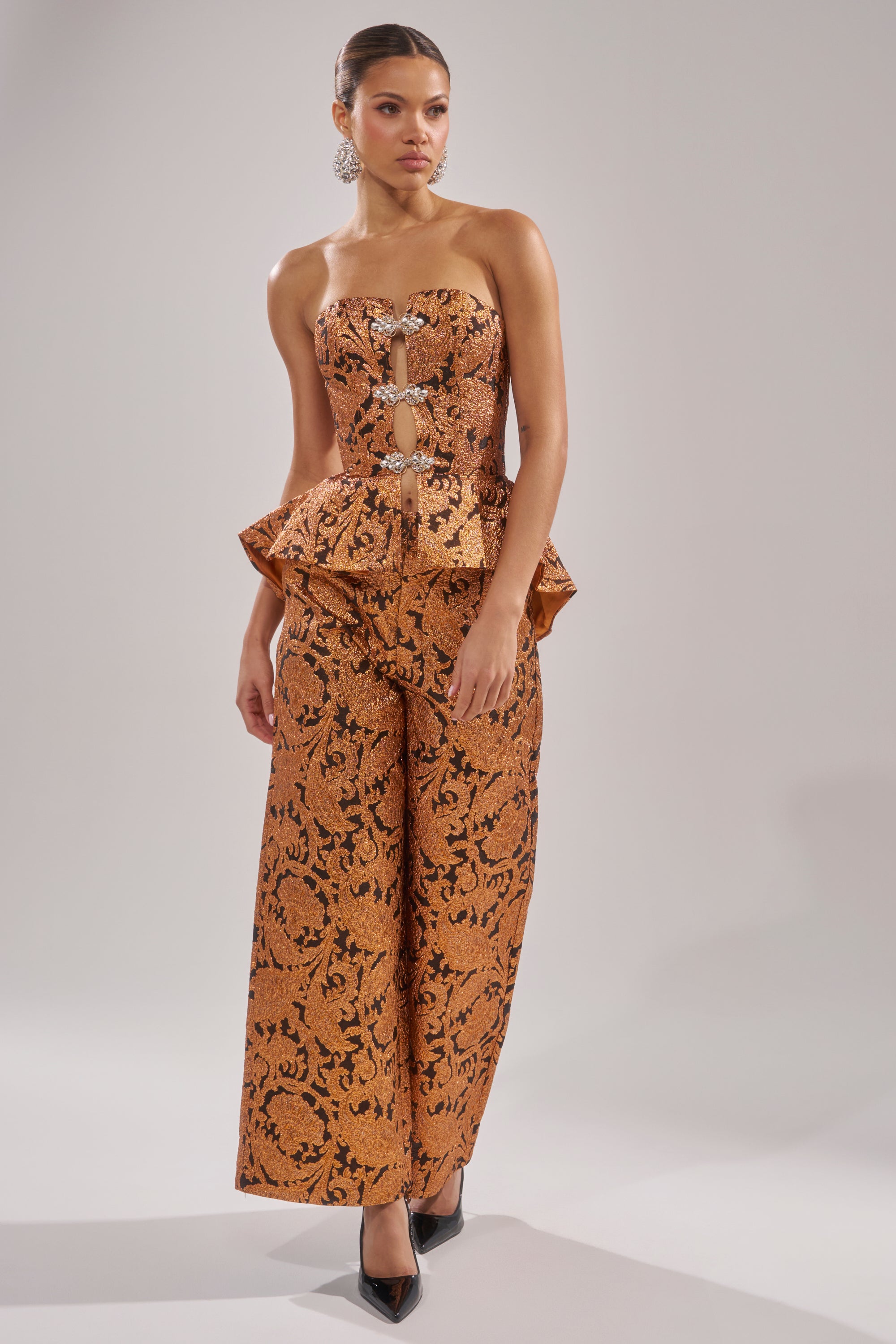 A woman models the AWAY FROM ME BROCADE TROUSER, a copper strapless jumpsuit with black floral brocade, peplum waist, front cutouts, and wide-leg silhouette. She pairs it with black pointed heels against a plain light background.