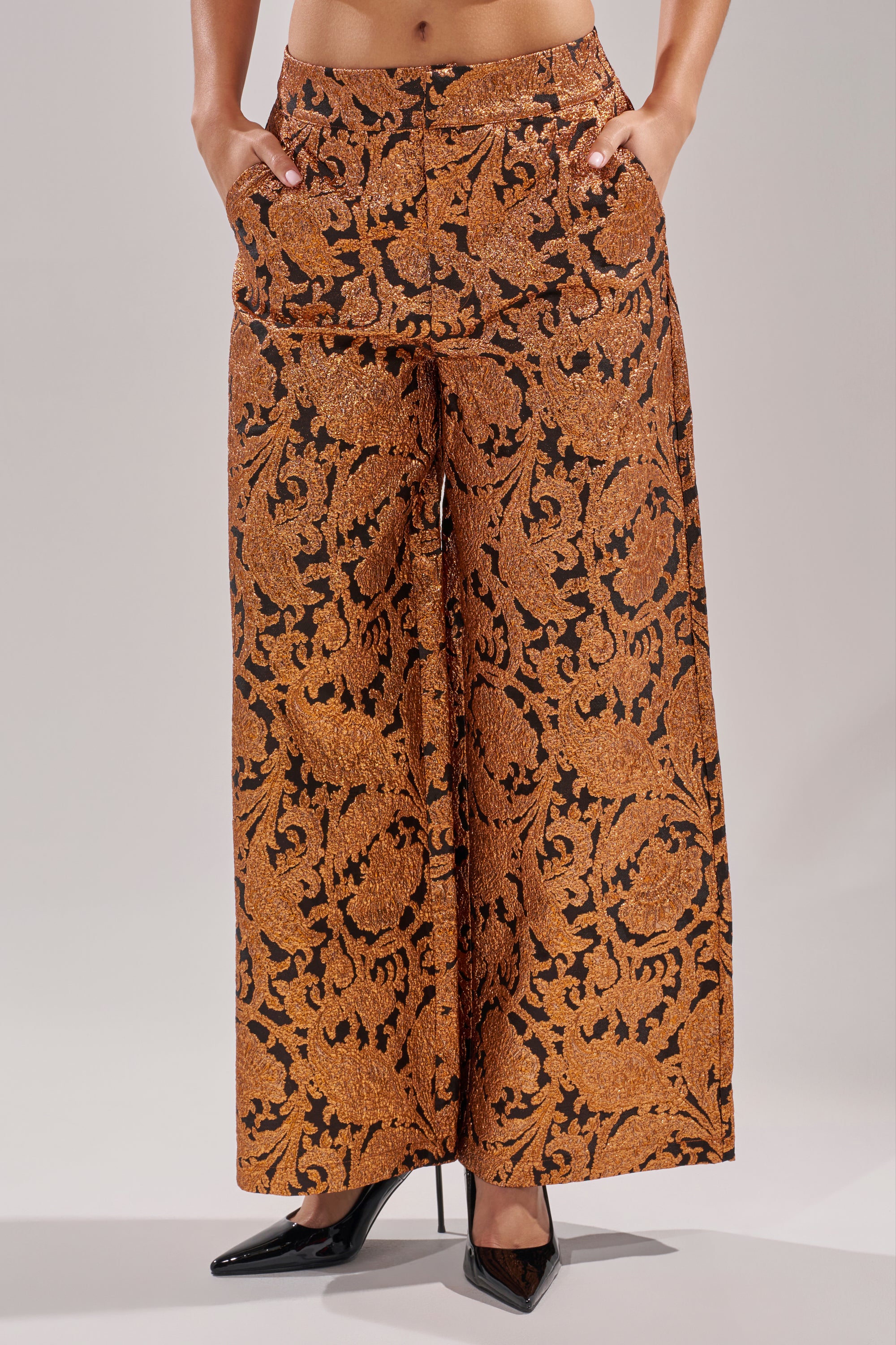 Wearing the AWAY FROM ME BROCADE TROUSER featuring a high waist, wide-leg silhouette, and a brown and black ornate floral pattern, styled with hands in pockets and finished with black pointed-toe heels.
