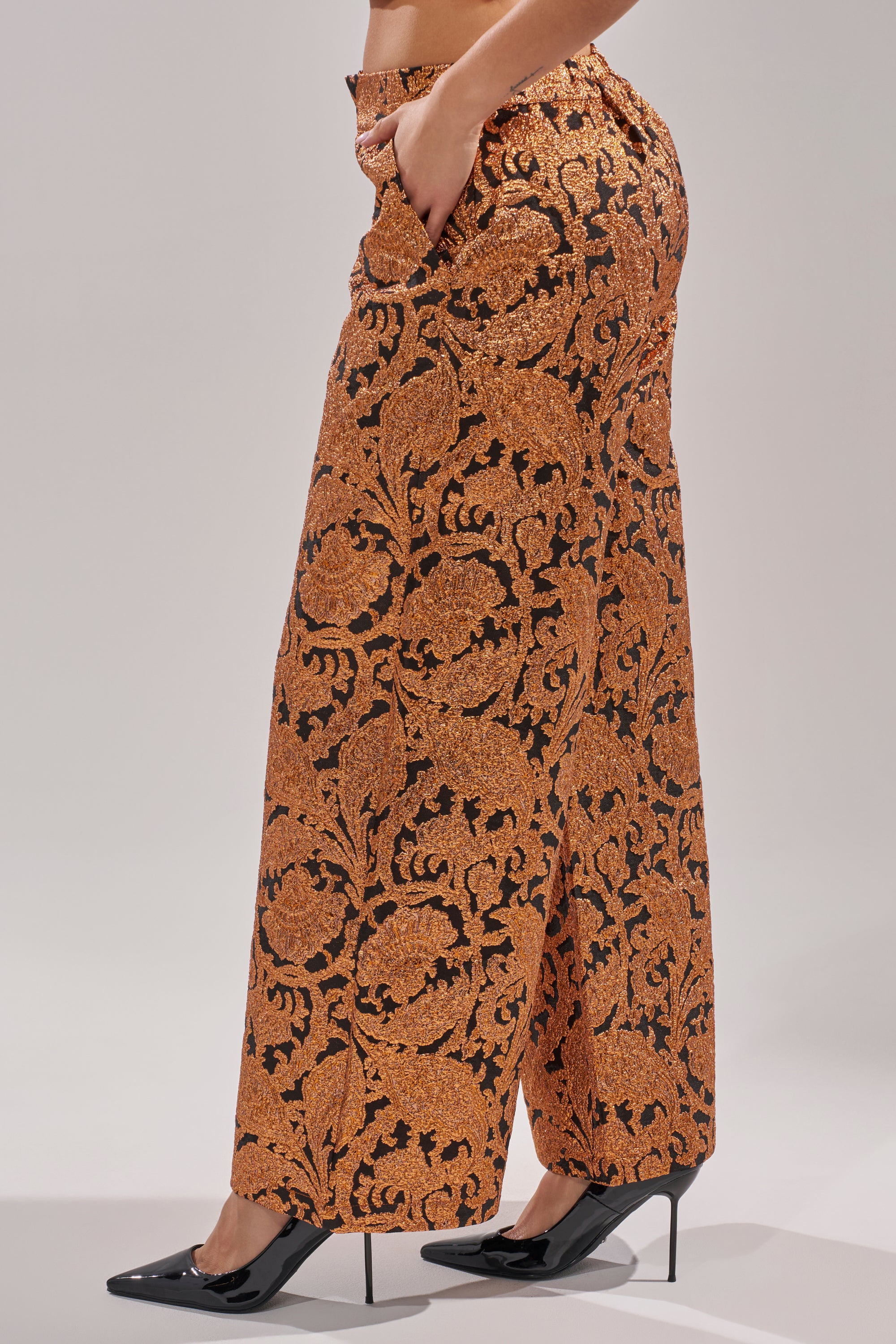 A person stands against a plain light background wearing the AWAY FROM ME BROCADE TROUSER—high-waisted, wide-leg pants with an orange and black floral pattern—paired with black pointed-toe heels.