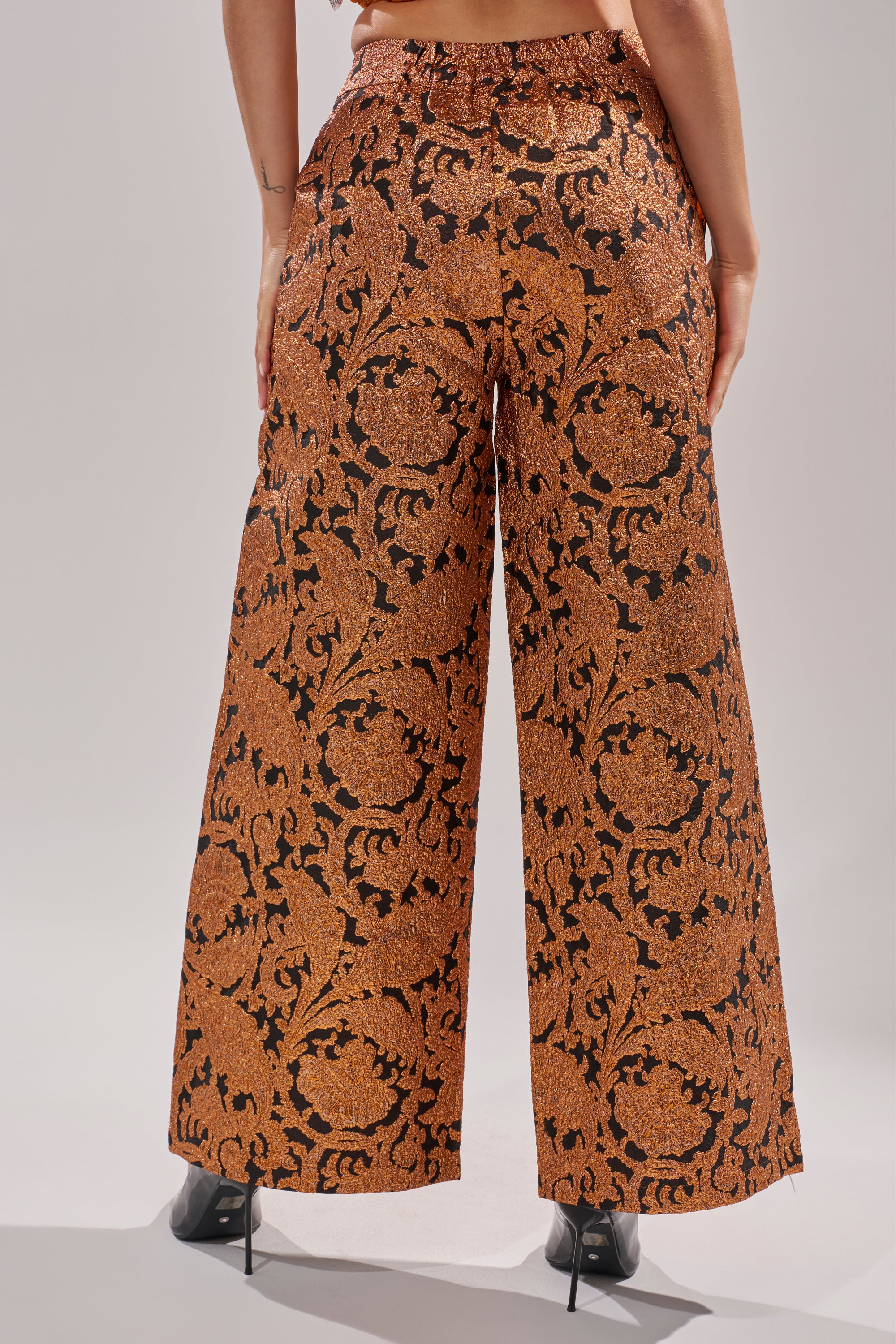 A person stands facing away, wearing the AWAY FROM ME BROCADE TROUSER featuring an ornate gold and black baroque pattern, paired with black high-heeled shoes against a plain, light-colored background.