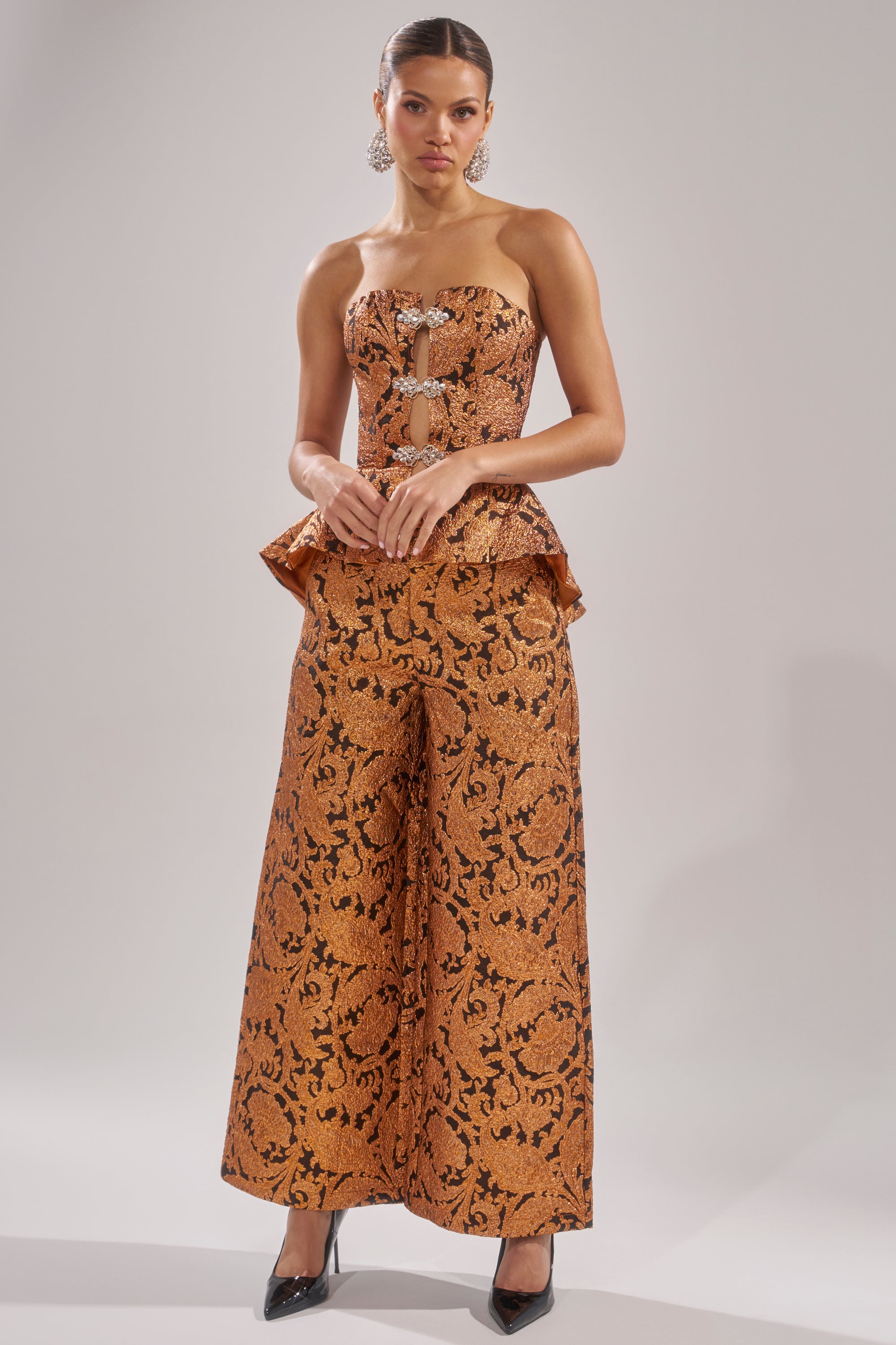 A woman poses in the AWAY FROM ME BROCADE TROUSER, a high-waisted, wide-leg silhouette pant with an orange and black brocade pattern, paired with a matching strapless peplum top, black heels, and statement earrings against a light grey background.