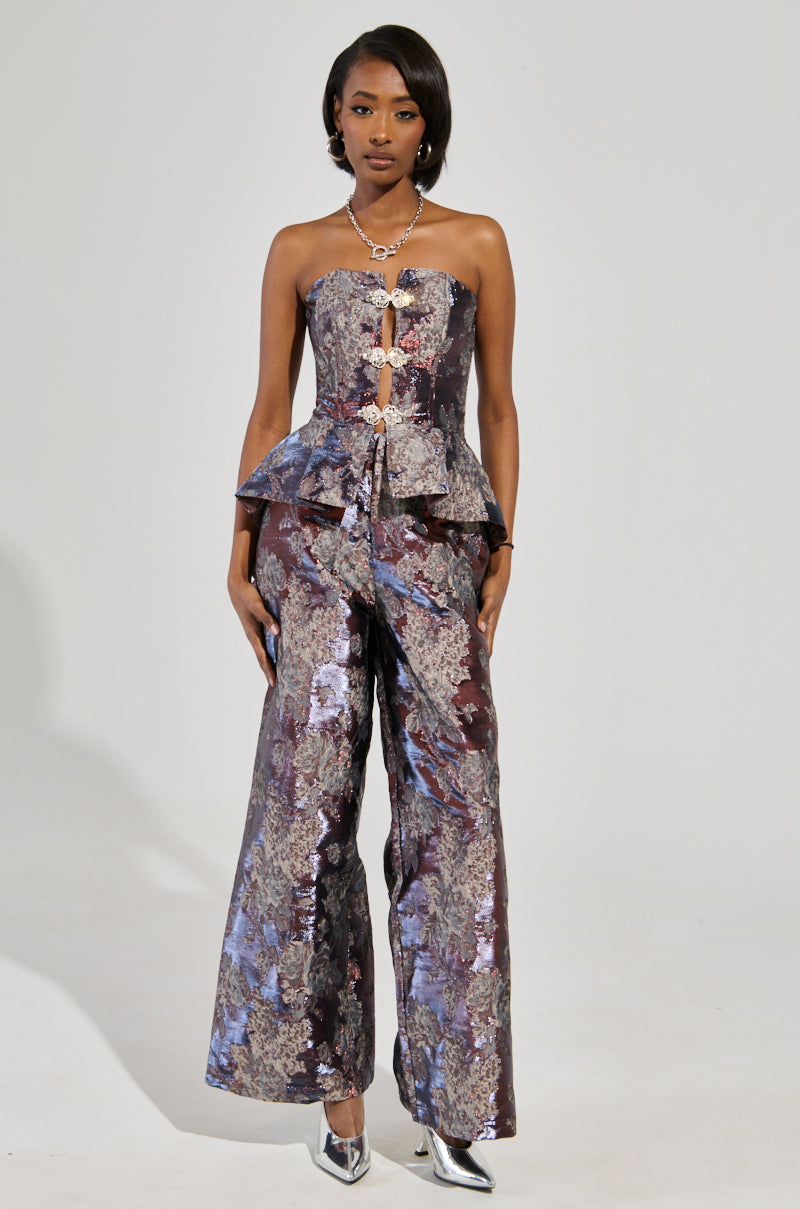 A person stands confidently in a stylish, strapless peplum top featuring an abstract floral brocade design and paired with the elegant ODESSA BROCADE TROUSER. The ensemble showcases shimmering metallic tones, while silver heels and a statement necklace add the perfect finishing touches.