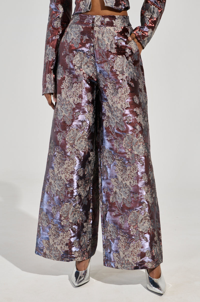 Dressed in the striking ODESSA BROCADE TROUSER, featuring a wide-leg design and a luxurious floral brocade pattern in purple and silver, the individual showcases a matching top with visible sleeves. The outfit is elegantly completed with metallic silver shoes, creating a stunning contrast against a plain white background.