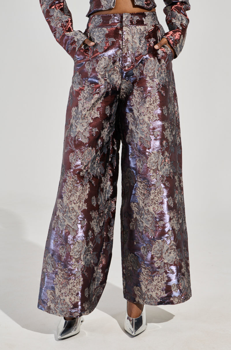 A person is captured wearing the ODESSA BROCADE TROUSER, featuring a dazzling high-waisted design and stunning floral brocade pattern. These wide-legged pants are beautifully complemented by pointed metallic shoes, creating a chic and modern look. The focus on just the lower half adds a sense of mystery.
