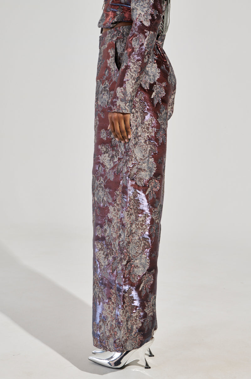 A person is dressed in a long metallic ensemble featuring floral brocade patterns in burgundy and silver hues. The form-fitting outfit reaches the ground, paired elegantly with shiny, silver pointed high-heeled boots for a captivating look.