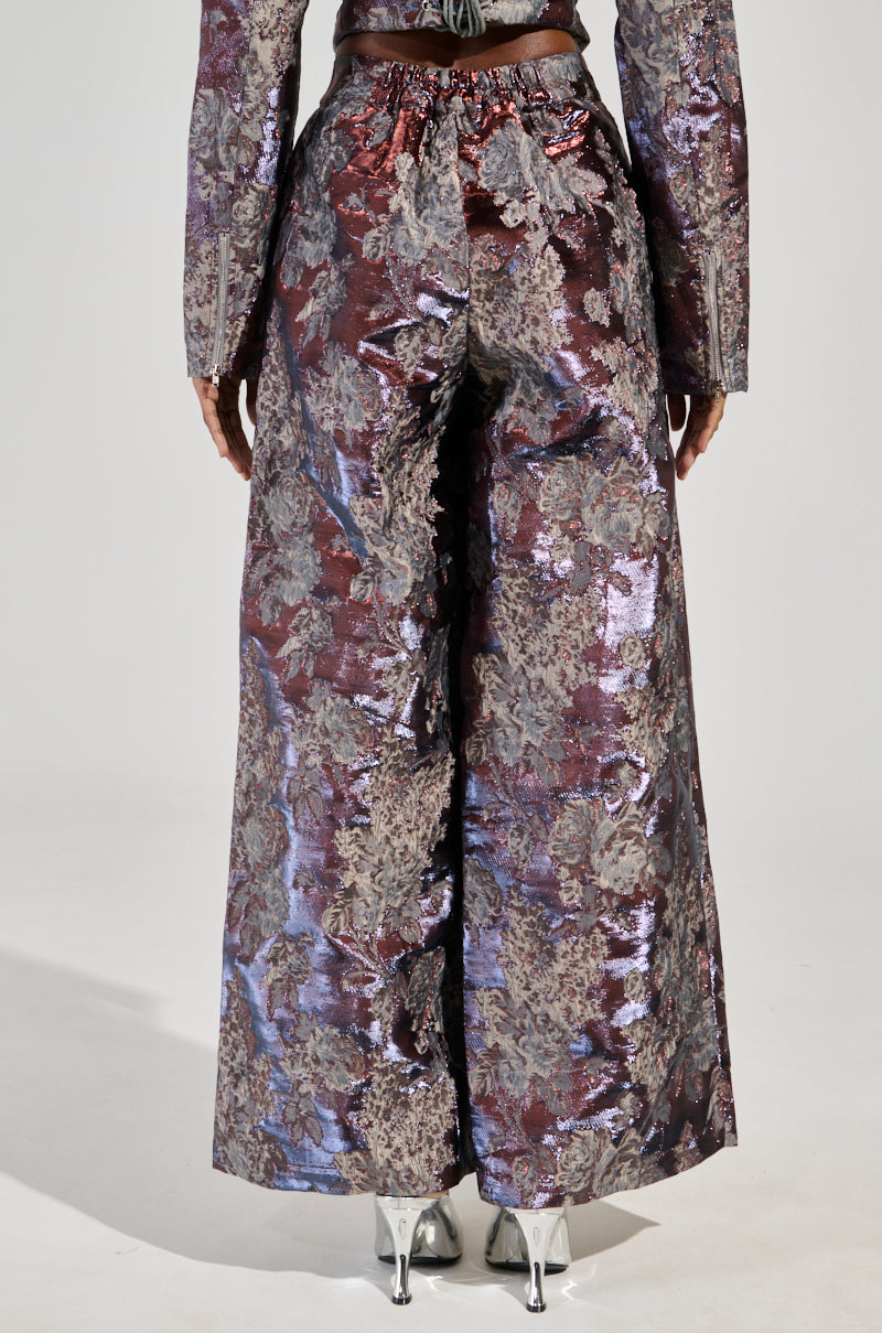 A person is dressed in the ODESSA BROCADE TROUSER, featuring a wide-leg silhouette crafted from metallic jacquard with a floral brocade design in purple and silver. Paired with a coordinating long-sleeve top and sleek silver shoes, the individual is photographed from behind against a simple backdrop.