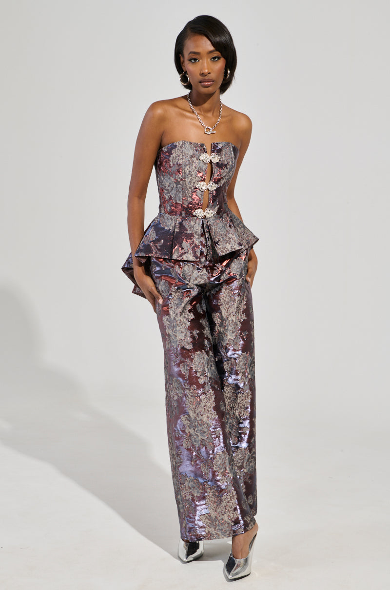 A woman showcases the ODESSA BROCADE TROUSER, pairing it with a strapless jumpsuit in silver and maroon floral brocade. The ensemble features a peplum waist and decorative buttons. She completes her look with silver heels and layered necklaces while posing against a plain gray backdrop.