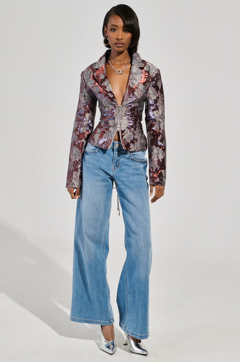 A person poses in a fashionable ensemble, featuring the ODESSA BROCADE BLAZER with a floral design and wide-leg blue jeans. They complement their look with metallic, color-shifting silver heels. Their hair is sleekly styled, and they are accessorized with a necklace against a plain white backdrop.