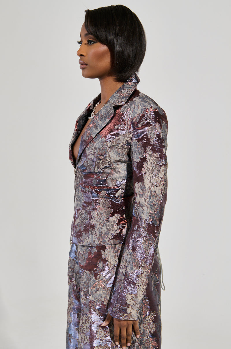 A person wearing the ODESSA BROCADE BLAZER stands in profile against a plain background, showcasing its metallic color-shifting textures with floral patterns in shades of purple and silver. The individual has short, sleek hair.