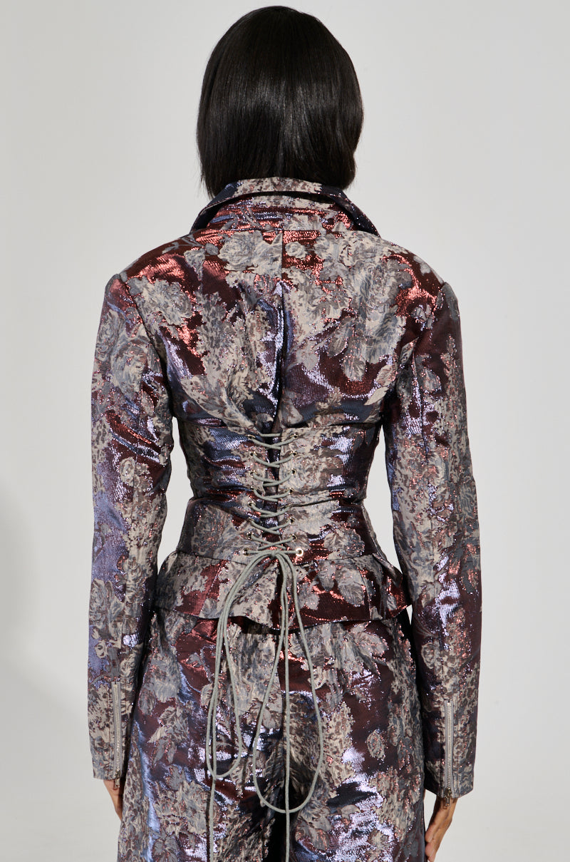 A person with long dark hair is wearing the ODESSA BROCADE BLAZER, which features a corset-style back with intricate lacing. The photograph captures them from behind against a plain background, emphasizing the blazer's metallic color-shifting patterns.