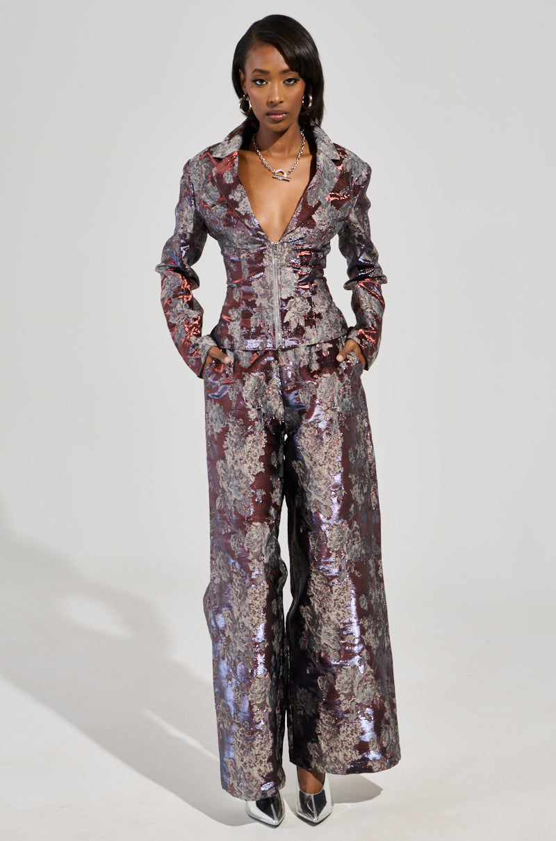 A person is wearing the Odessa Brocade Blazer with matching wide-leg pants, featuring a shiny, floral brocade pattern and a deep V-neckline. The metallic, color-shifting outfit beautifully blends shades of silver and burgundy. They stand confidently with hands in pockets, complemented by pointed silver shoes.