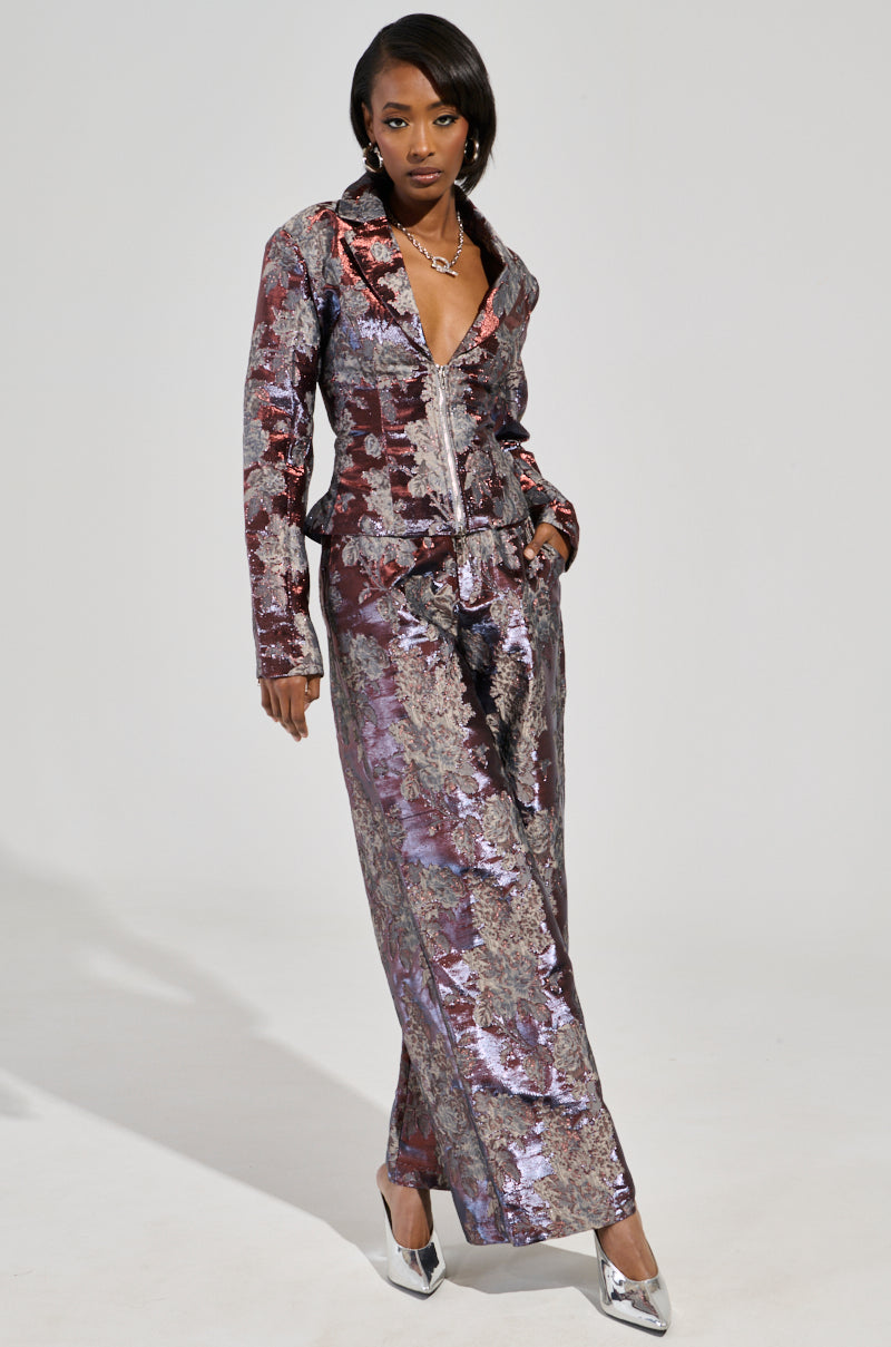 A woman poses confidently in the stylish ODESSA BROCADE BLAZER, featuring a silver-and-purple brocade design with a deep V-neck, paired with wide-legged pants. Accentuating her floral brocade pattern ensemble, she complements it with metallic pointed-toe heels. The plain background highlights her striking attire.