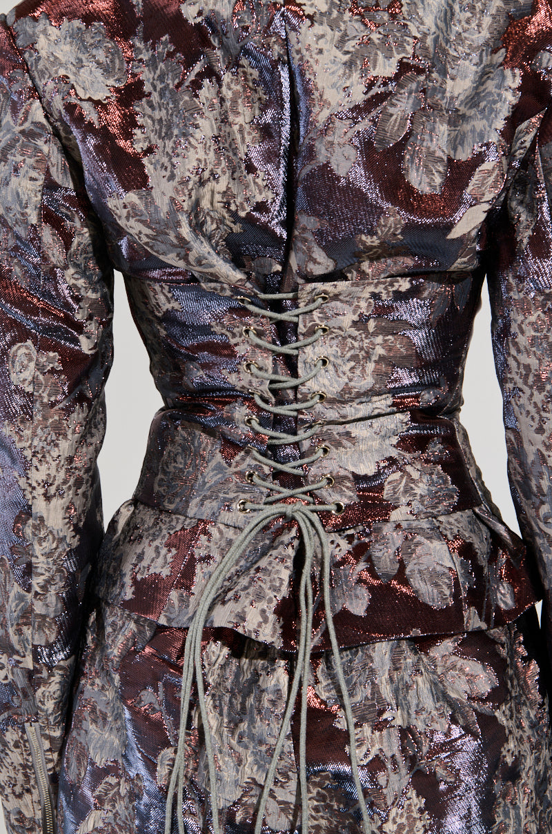 A detailed view of the ODESSA BROCADE BLAZER showcases its exquisite metallic color-shifting fabric with a corset-style back. The material glistens with a floral pattern in hues of purple, blue, and touches of brown. The laces are elegantly tied at the center, enhancing its intricate appeal.