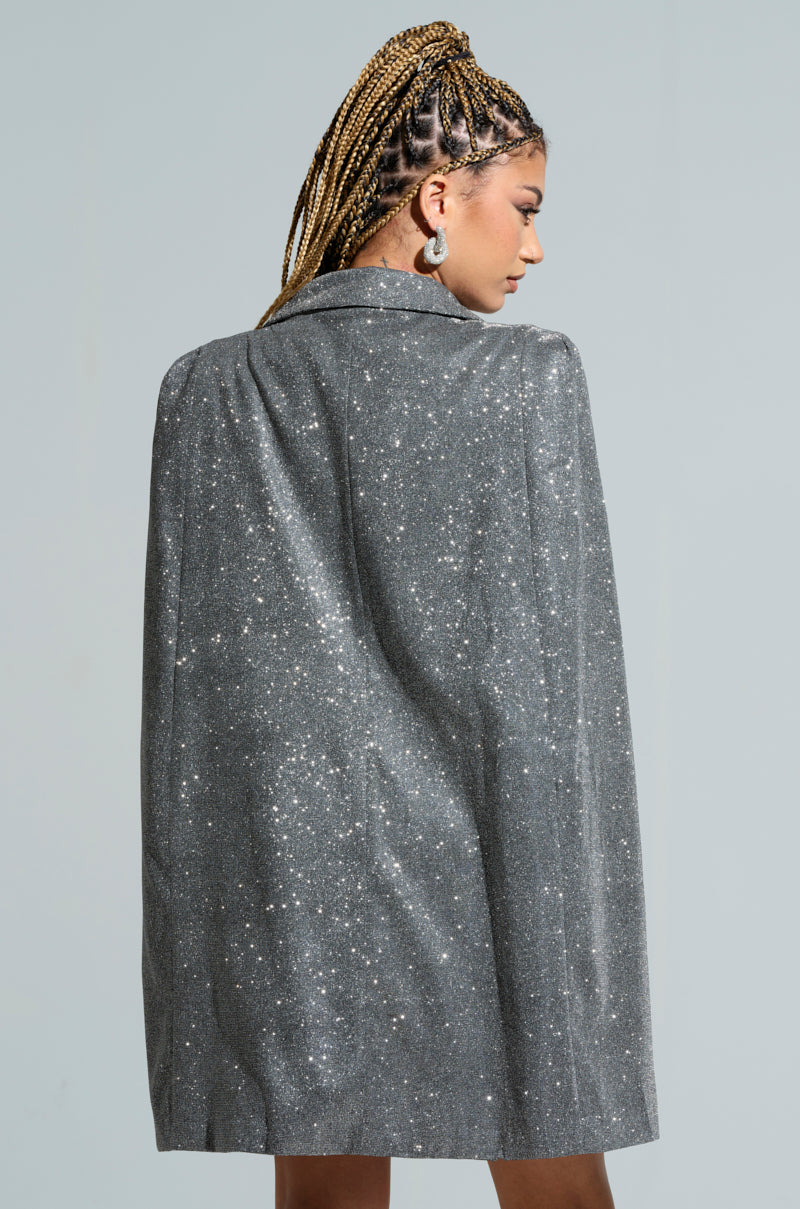 A woman with braided hair stands in profile, wearing the ALL ABOUT ME SPACESHINE BLAZER DRESS, a shimmering silver cape blazer dress with a plunging V-neckline. She is accessorized with large hoop earrings against a light blue background, showcasing perfect holiday fashion.