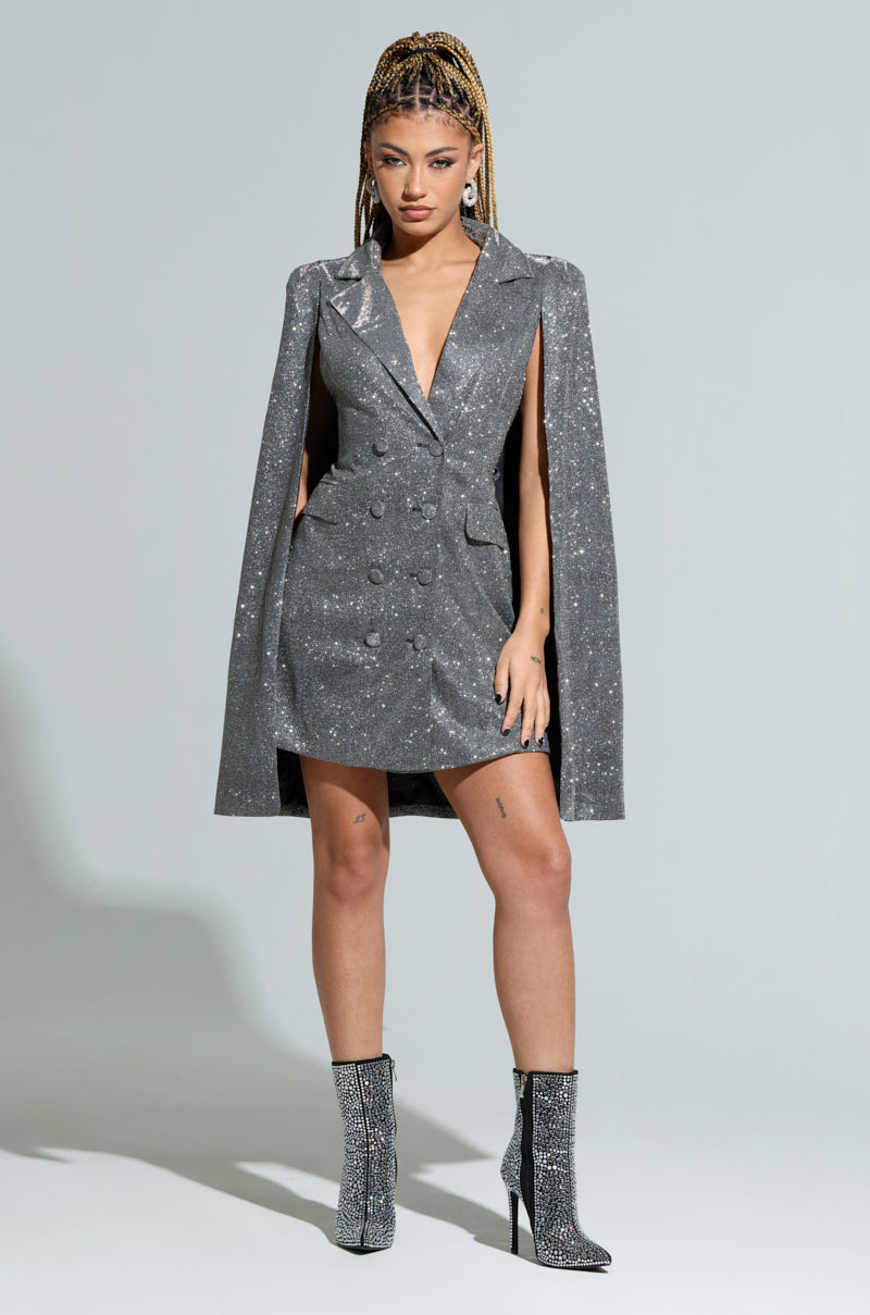 Against a gray backdrop, a person exudes holiday fashion in the ALL ABOUT ME SPACESHINE BLAZER DRESS, featuring cape-style sleeves and matching high-heeled boots. With long, braided hair styled in an updo and a confident expression, they embrace the spotlight with elegance.