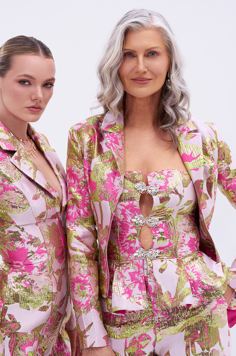 Two women wear LOVELY FLORAL CORSET suits in pink, gold, and green. One pairs it with a cutout top and long gray waves; the other styles a high-low blazer with slicked-back blonde hair. Both confidently face the camera.