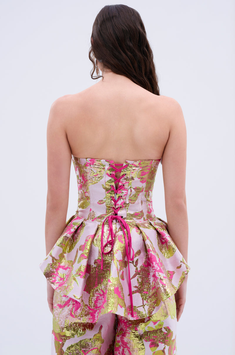 A woman with long wavy brown hair is seen from behind wearing the LOVELY FLORAL CORSET, featuring pink lace-up details, a peplum waist, and gold, pink, and green floral patterns, paired with matching pants for a party-ready look.