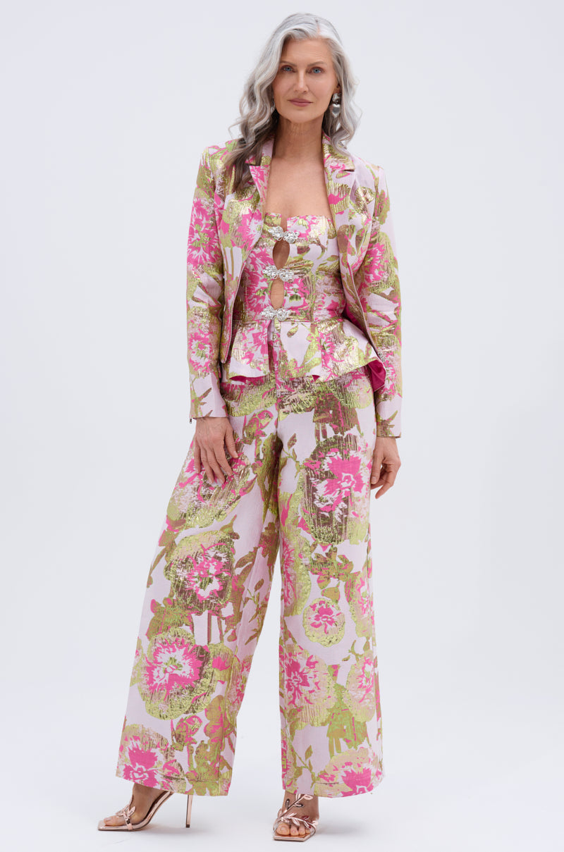 A woman with long, wavy gray hair stands confidently in open-toe heels against a plain background, wearing the LOVELY FLORAL CORSET and a floral suit with a fitted blazer, wide-leg pants, and pink, green, and gold cutouts.