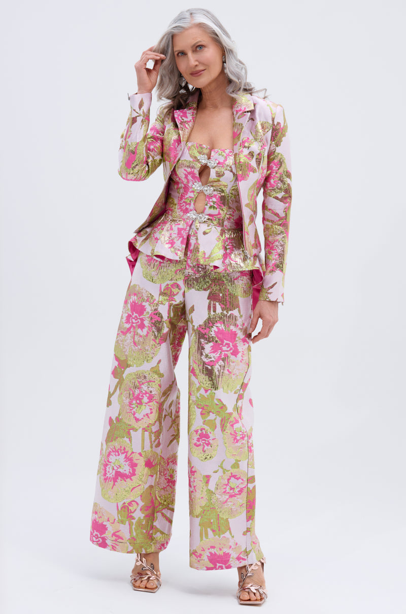 A woman with long gray hair wears the LOVELY FLORAL CORSET, styled with wide-leg floral pants and a matching fitted blazer in pink, green, and gold, plus metallic sandals. She stands against a plain white background.