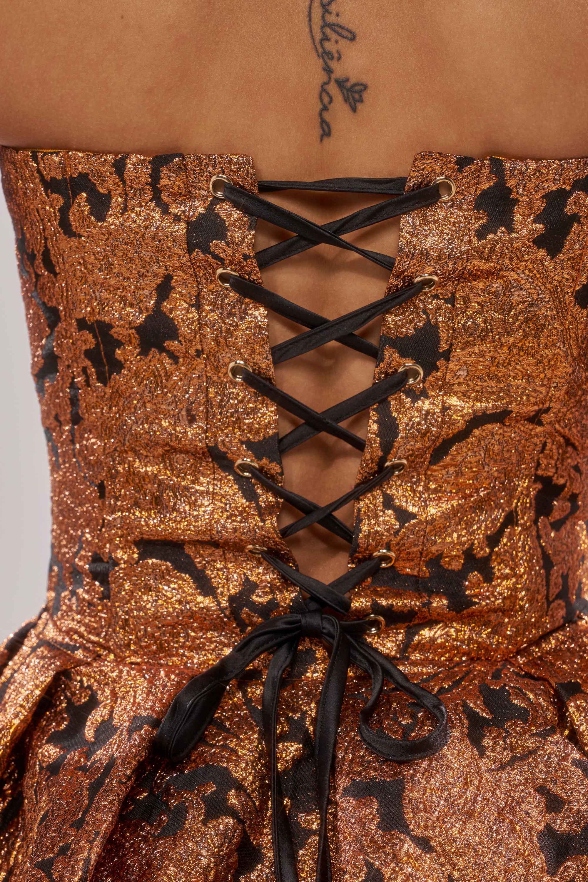 Close-up of the AWAY FROM ME BROCADE CORSET in metallic bronze and black, featuring brocade fabric, a black lace-up ribbon back, and a visible upper back tattoo with cursive text and a leaf motif.