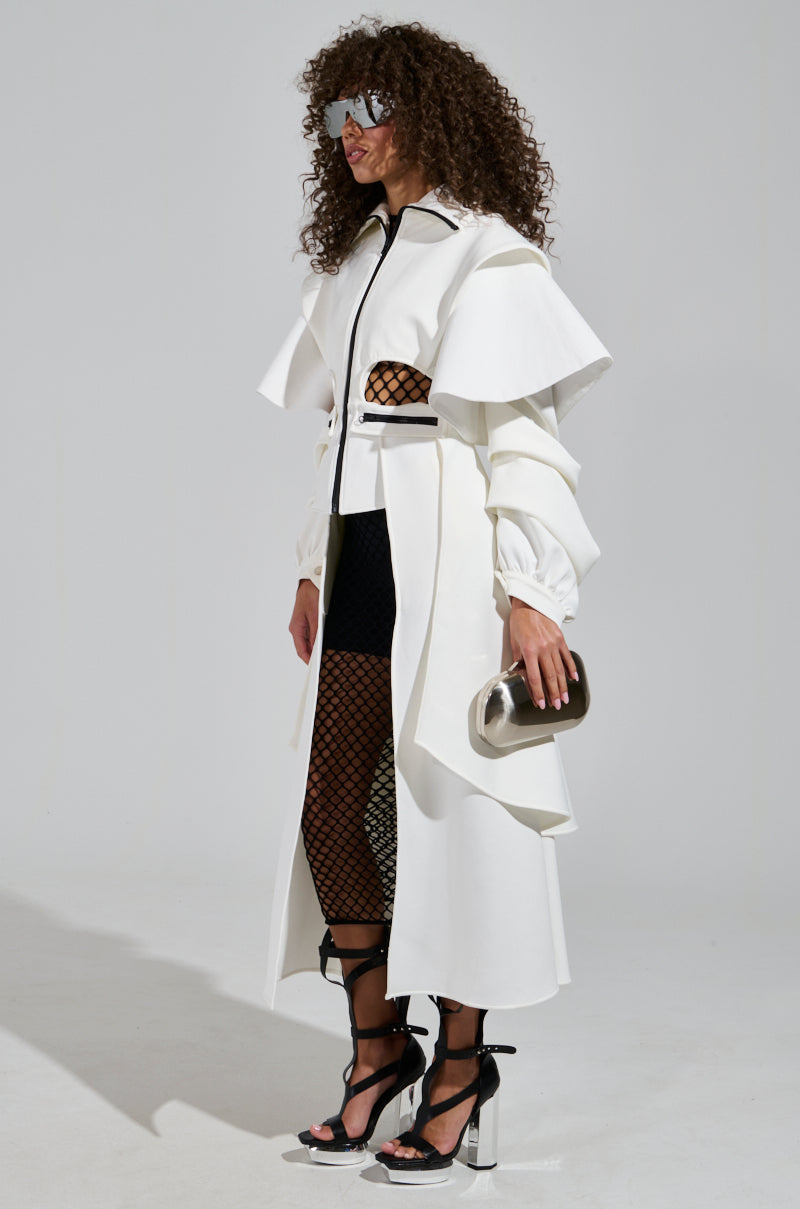 A person with curly hair exudes style in trendy outerwear featuring the FUTURA CUTOUT SCUBA TRENCH, a white coat with ruffles and a cape silhouette. They complement their look with black mesh underlay, sunglasses, and hold a metallic clutch while wearing high-heeled sandals with ankle straps against a plain background.