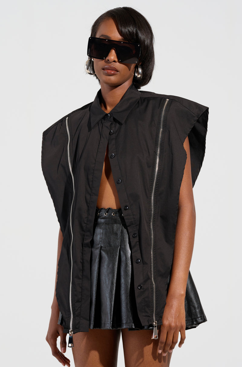 A person wearing oversized black sunglasses, the edgy-chic BAD GIRL ZIPPER DETAIL BUTTON DOWN SHIRT, and a pleated leather skirt poses confidently against a plain background.