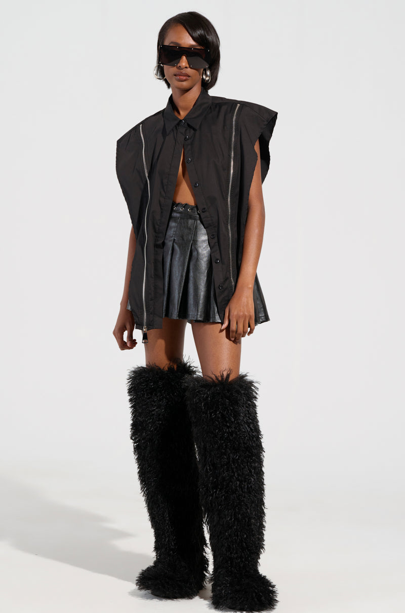 A person wearing oversized sunglasses, the BAD GIRL ZIPPER DETAIL BUTTON DOWN SHIRT, a short black leather skirt, and large furry black boots stands against a plain white background.