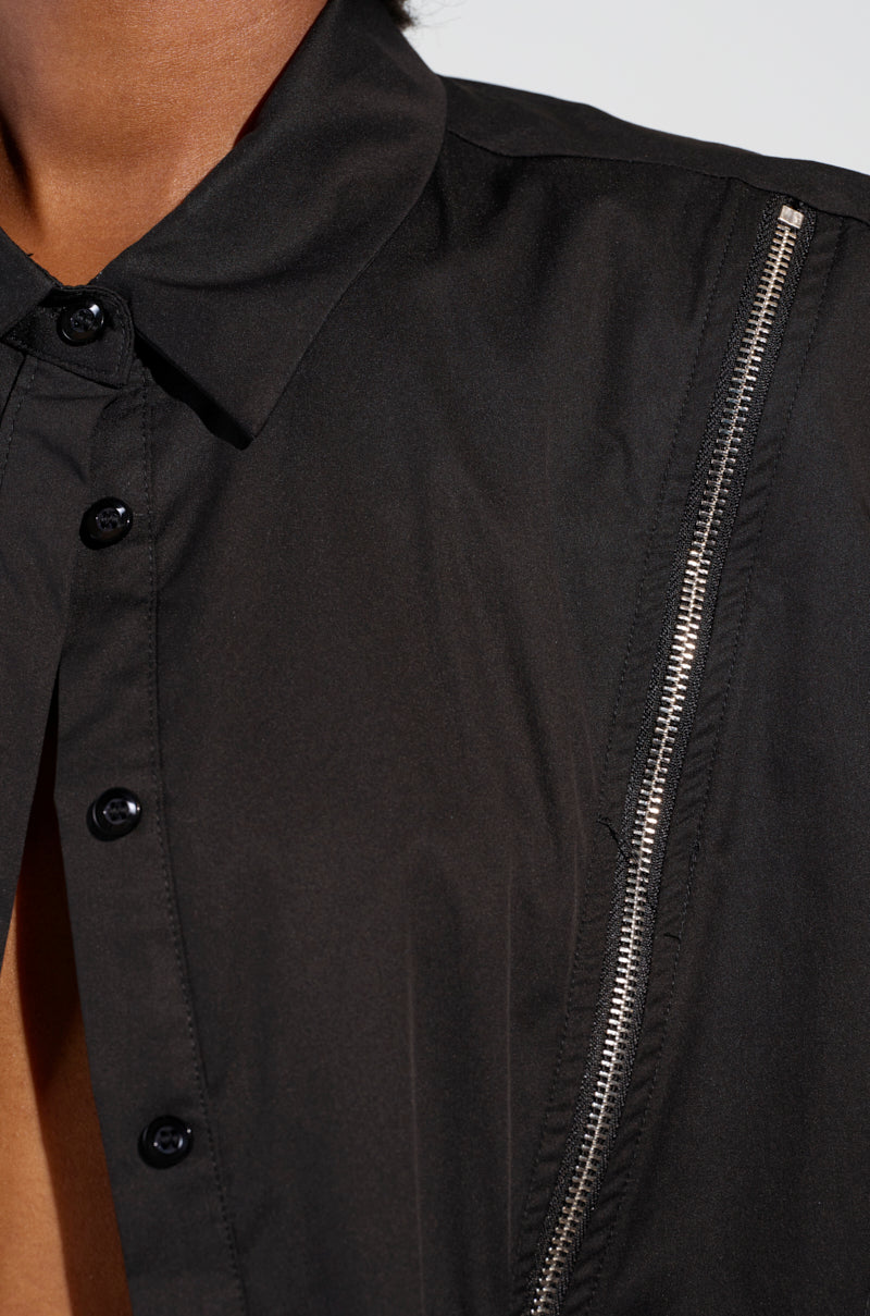 A close-up image shows someone wearing the BAD GIRL ZIPPER DETAIL BUTTON DOWN SHIRT, showcasing its edgy-chic style with a black button-up design. The shirt features metallic zipper detailing on the shoulder and is slightly open to reveal part of the chest. The smooth fabric and visible stitching add to its distinctive look.