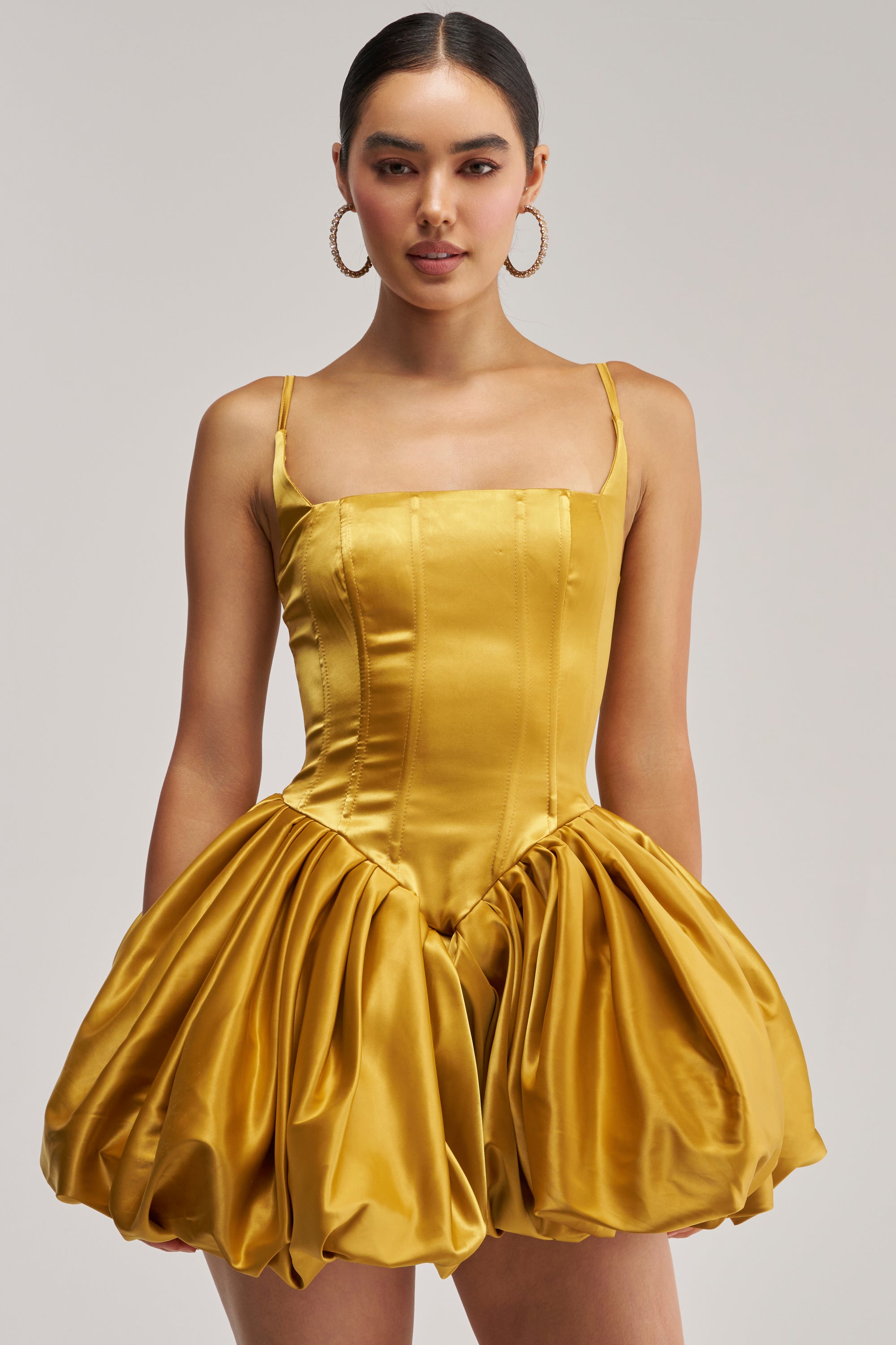 A woman stands against a plain light background, wearing the JULIET SATIN MINI DRESS IN MUSTARD, which features a fitted bodice and voluminous skirt. She accessorizes with hoop earrings and has her hair tied back.