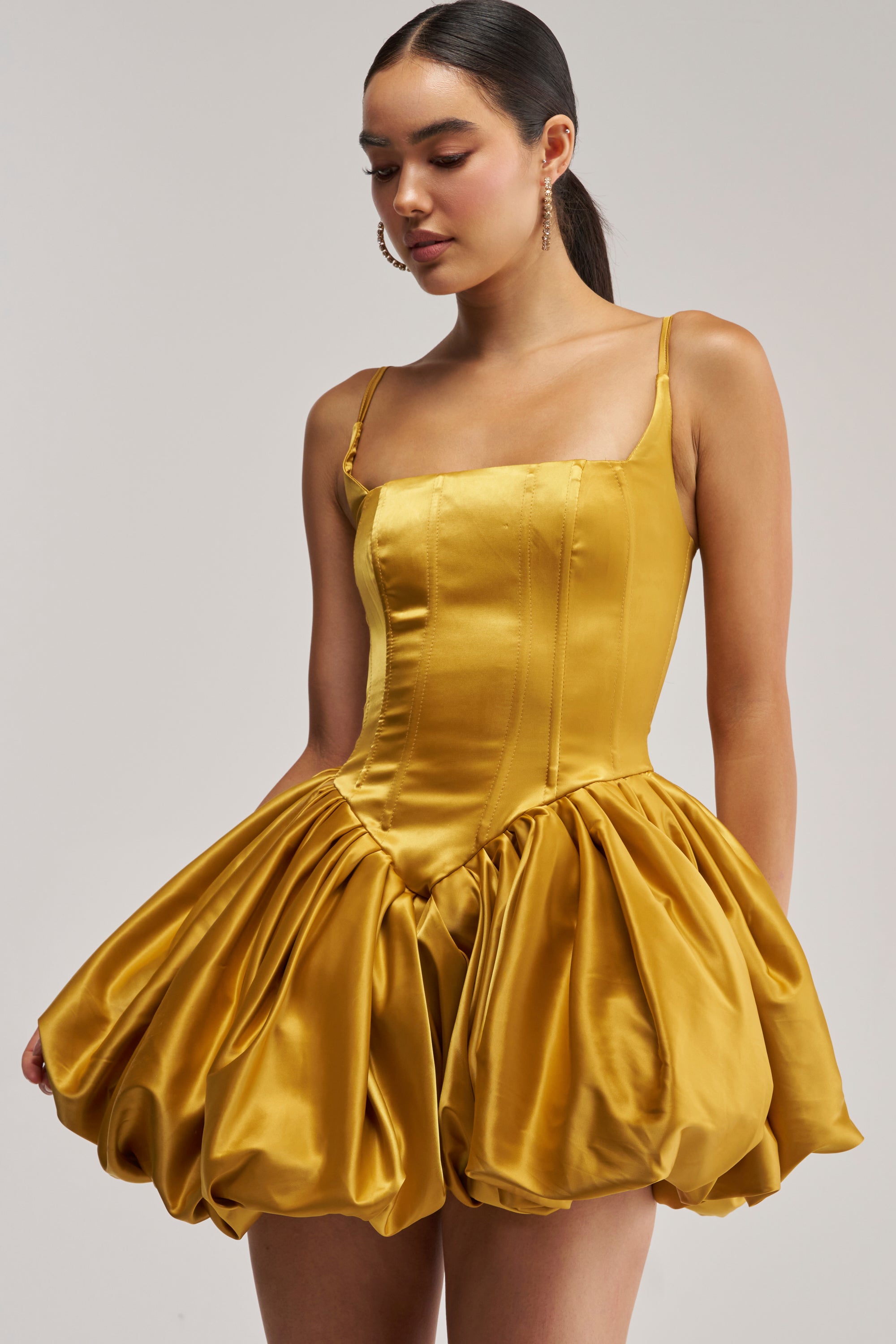 Wearing the JULIET Satin Mini Dress in Mustard, a woman looks down with her hair styled in a sleek ponytail; the shiny gold satin dress features a fitted bodice and a voluminous bubble skirt.