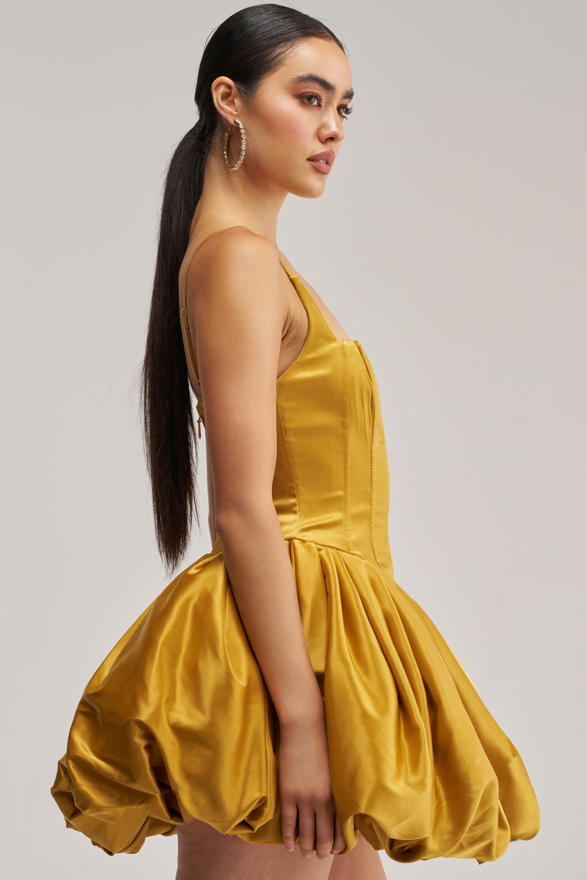 A woman in profile wears the JULIET SATIN MINI DRESS IN MUSTARD, a shiny mini dress with a fitted bodice and a voluminous bubble skirt. Her long, straight dark hair is styled in a low ponytail, and she accessorizes with large hoop earrings against a plain background—making this the perfect standout look for any homecoming moment.