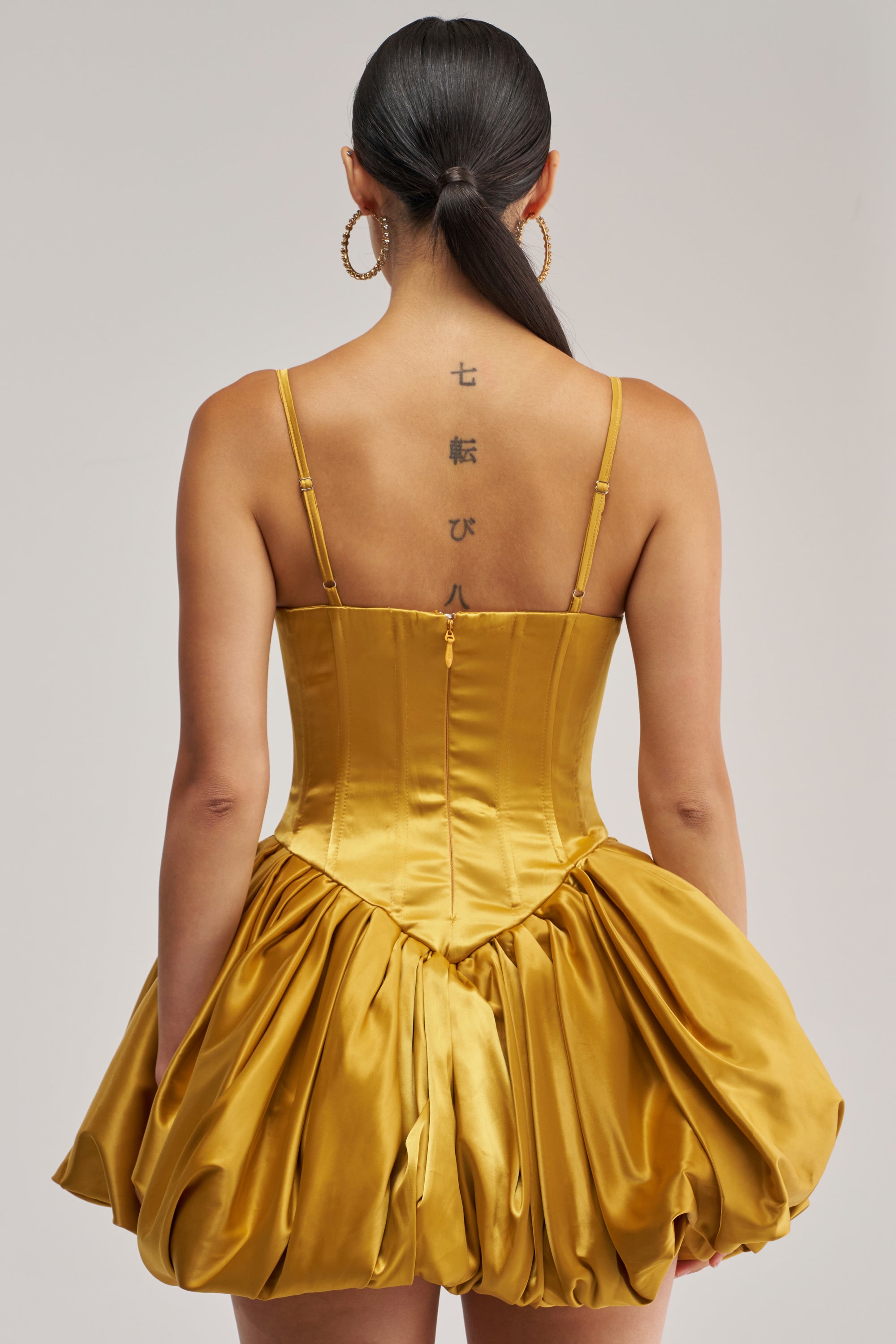 A woman with long dark hair in a ponytail stands with her back to the camera, showcasing a Japanese script tattoo along her spine and large hoop earrings. She is wearing the JULIET SATIN MINI DRESS IN MUSTARD, which features a shiny finish and a flared skirt.