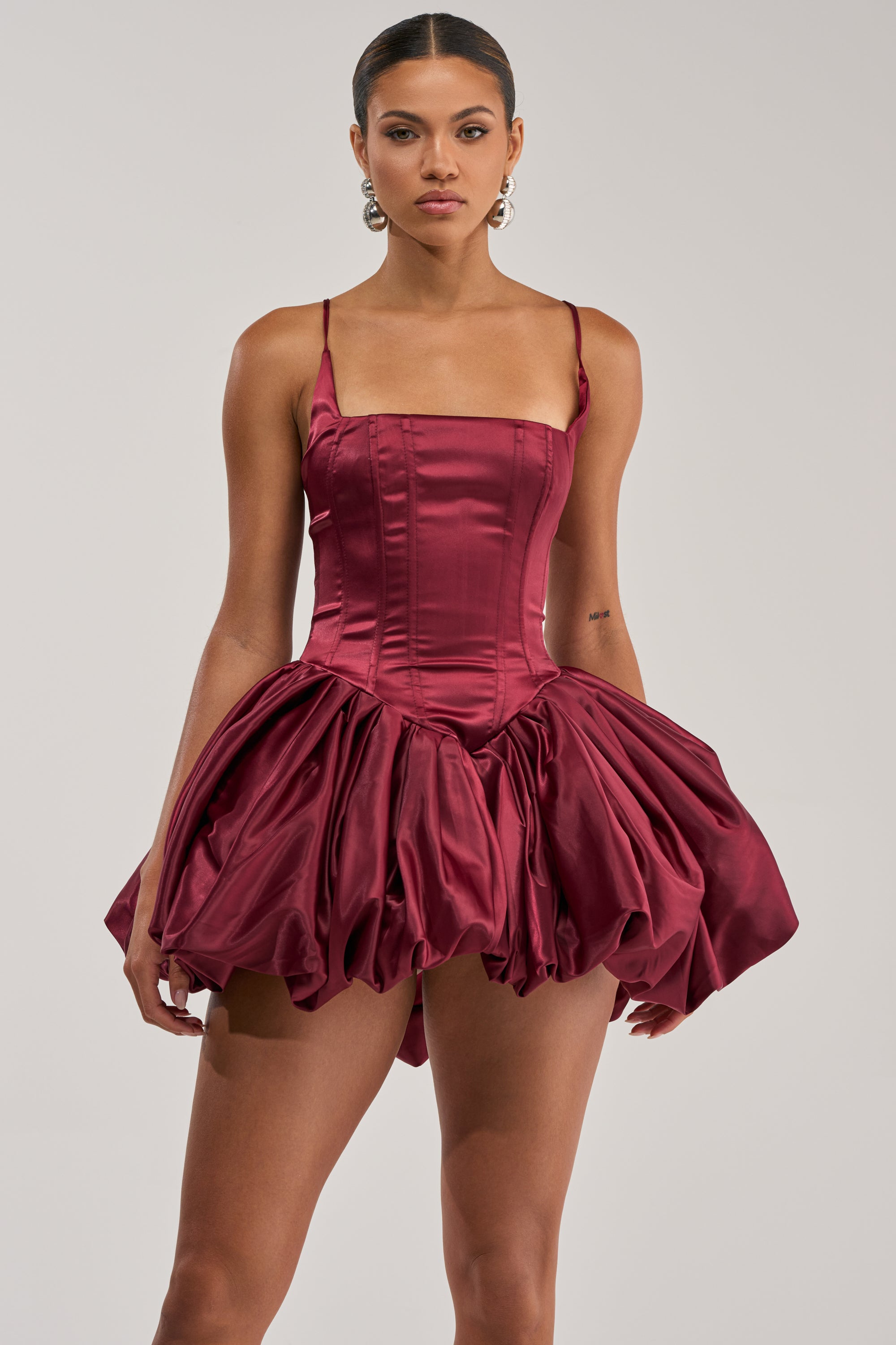 A woman models the JULIET SATIN MINI DRESS IN BURGUNDY, featuring a fitted bodice and voluminous bubble skirt. Against a light background, she wears hoop earrings and has her hair pulled back.