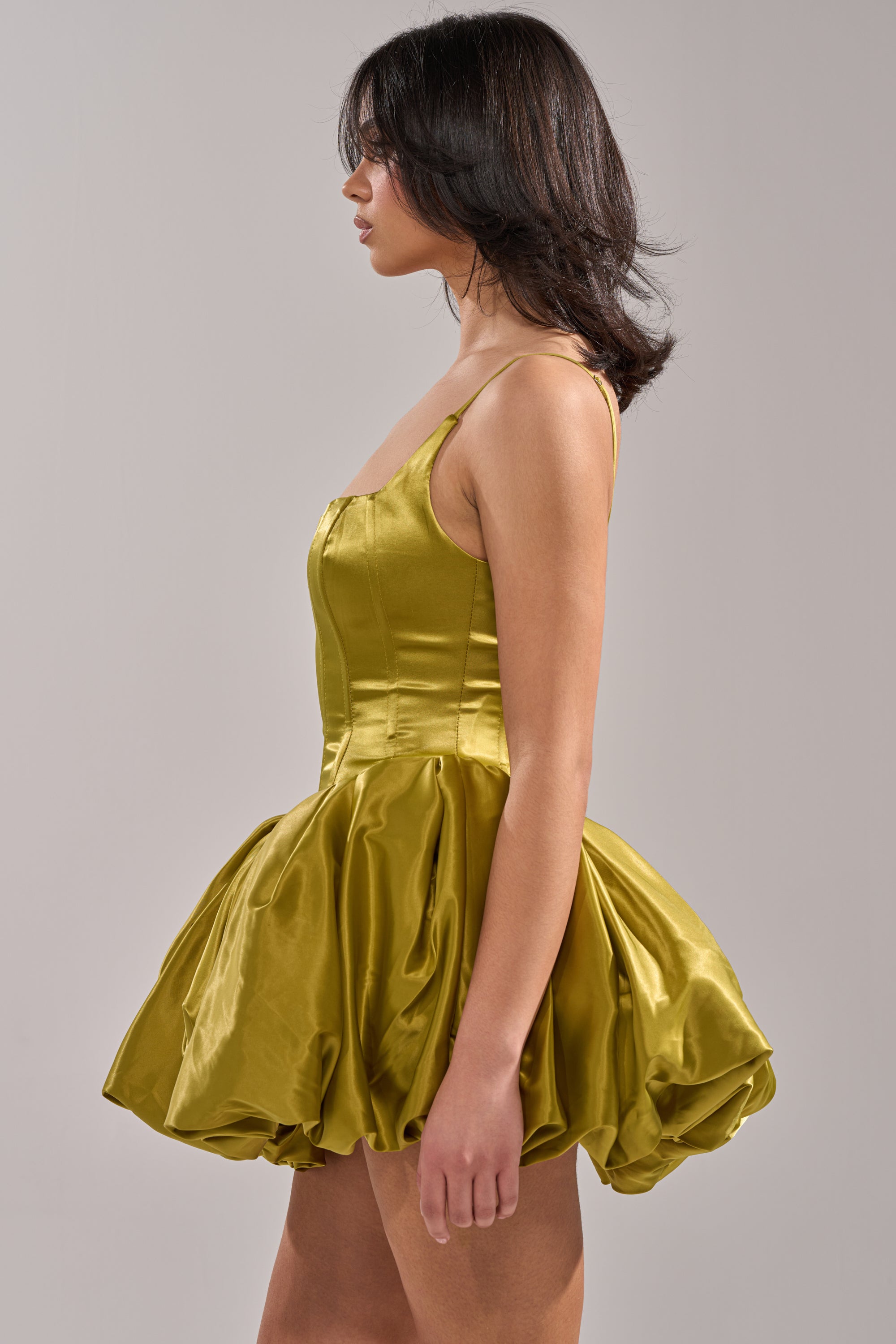 A woman stands in profile wearing the JULIET SATIN MINI DRESS IN GREEN, featuring a fitted bodice and voluminous bubble skirt. The plain, neutral background highlights the dress's unique design. 3