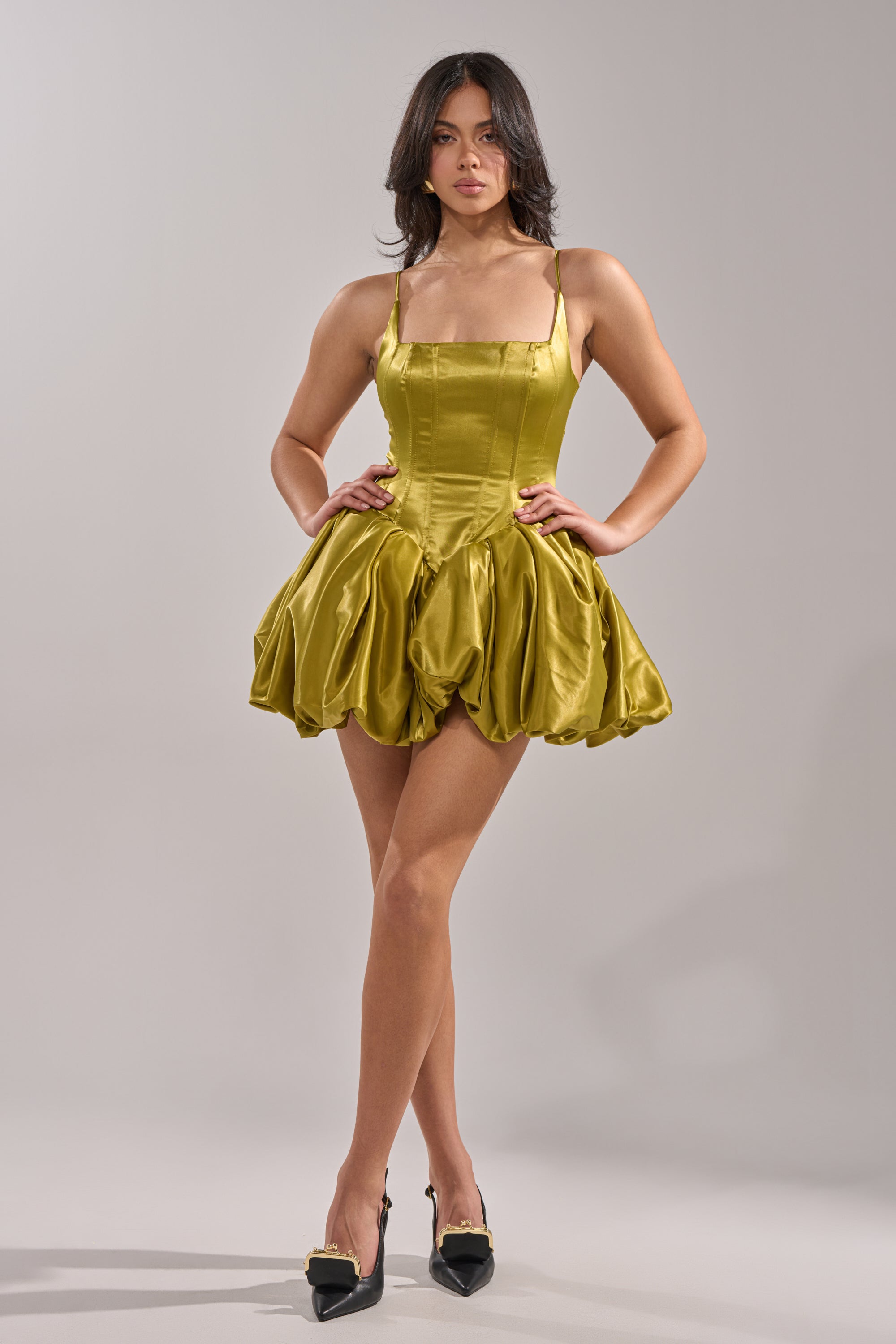 A woman stands confidently in the JULIET SATIN MINI DRESS IN GREEN, featuring a corset bodice and voluminous bubble skirt, styled with black pointed-toe heels, against a plain light gray background. 5