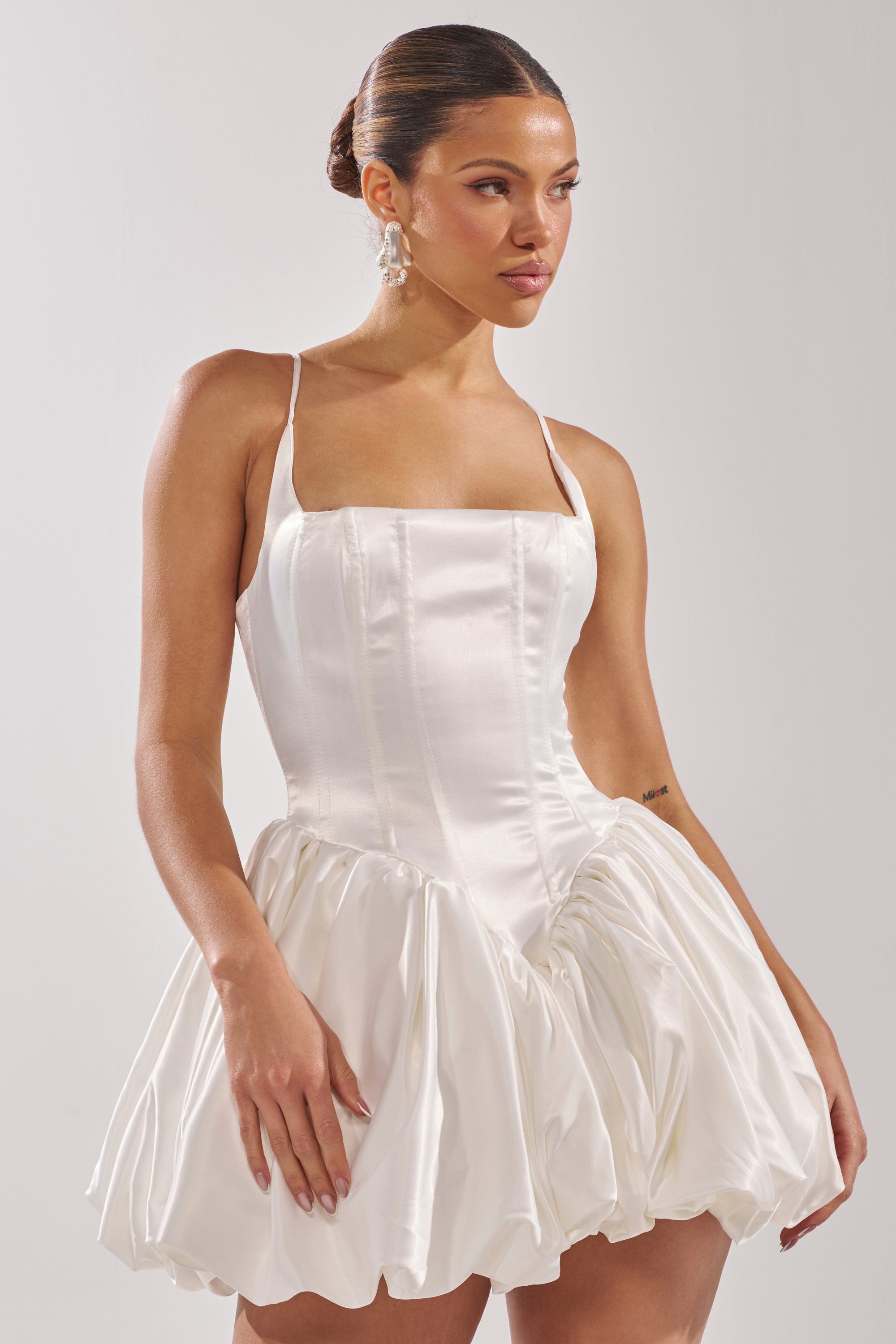 A woman wears the JULIET SATIN MINI DRESS IN IVORY, featuring a corset bodice, thin straps, and a voluminous bubble hem skirt. Her hair is styled in a sleek bun and she completes the look with dangling earrings.