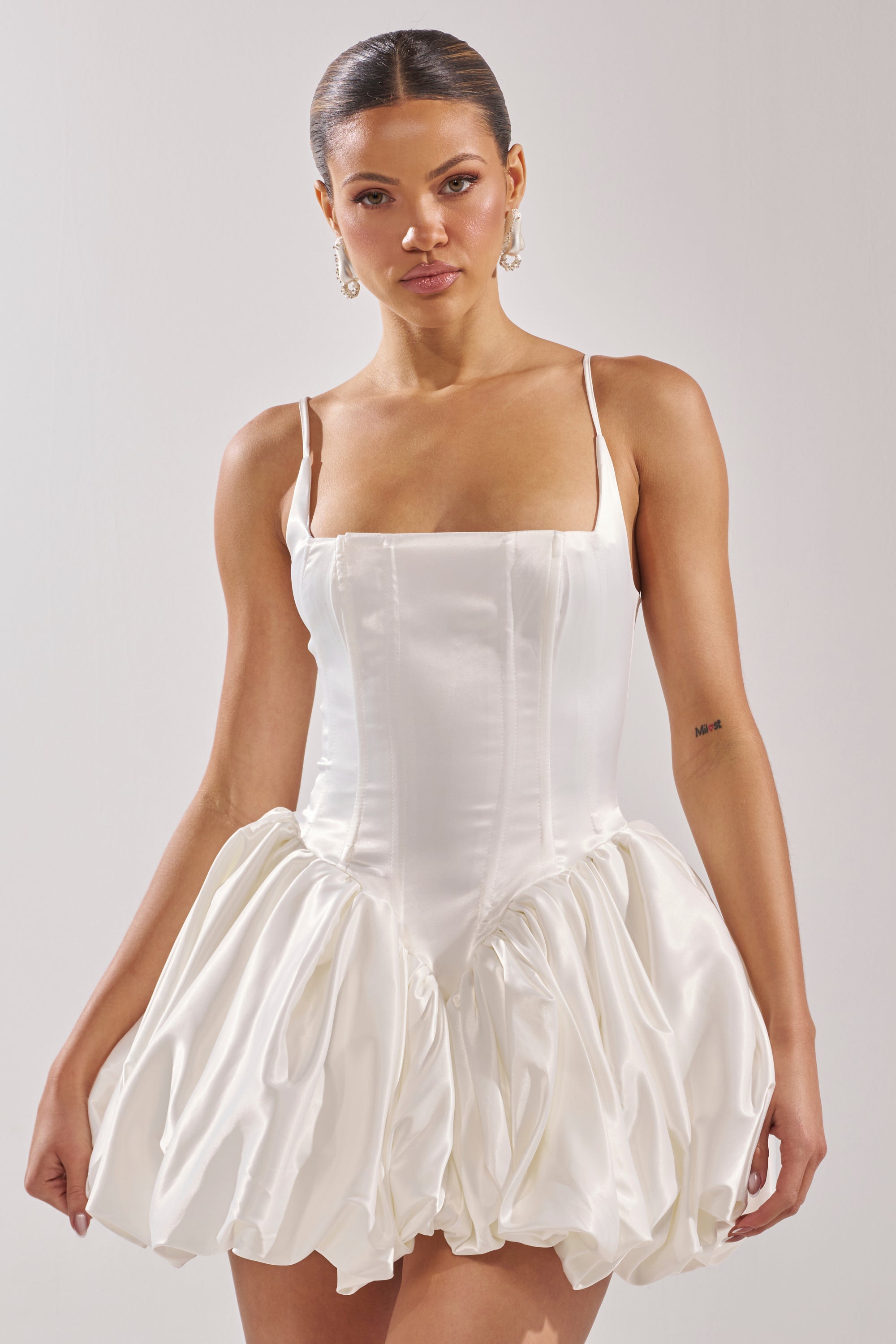 A woman models the JULIET SATIN MINI DRESS IN IVORY, featuring a corset bodice and voluminous bubble hem skirt. She stands against a plain light background with slicked-back hair and dangling earrings.