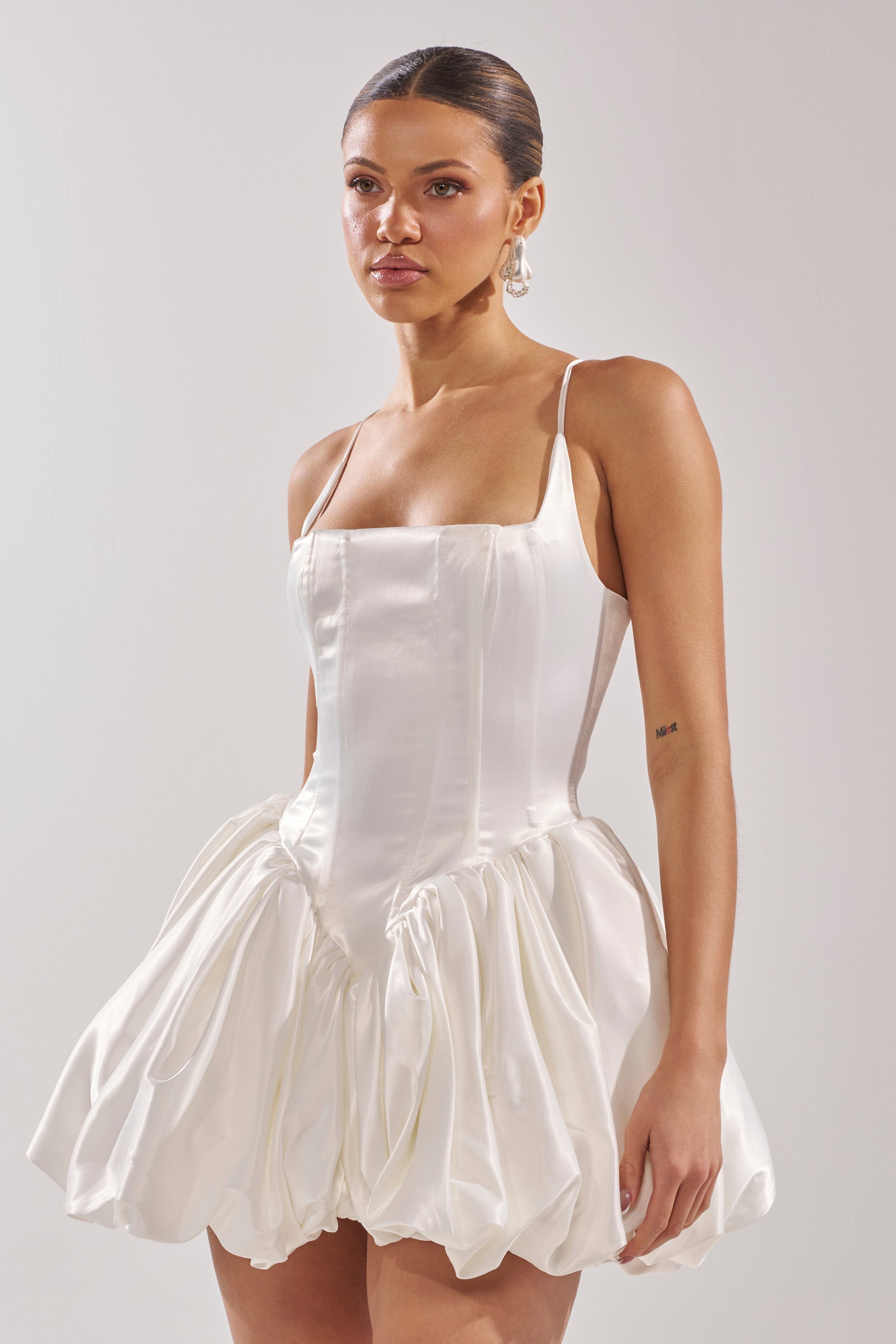 A woman wears the JULIET SATIN MINI DRESS IN IVORY, featuring a bubble hem skirt and spaghetti straps, against a plain light background. Her hair is slicked back and she finishes the look with elegant drop earrings.