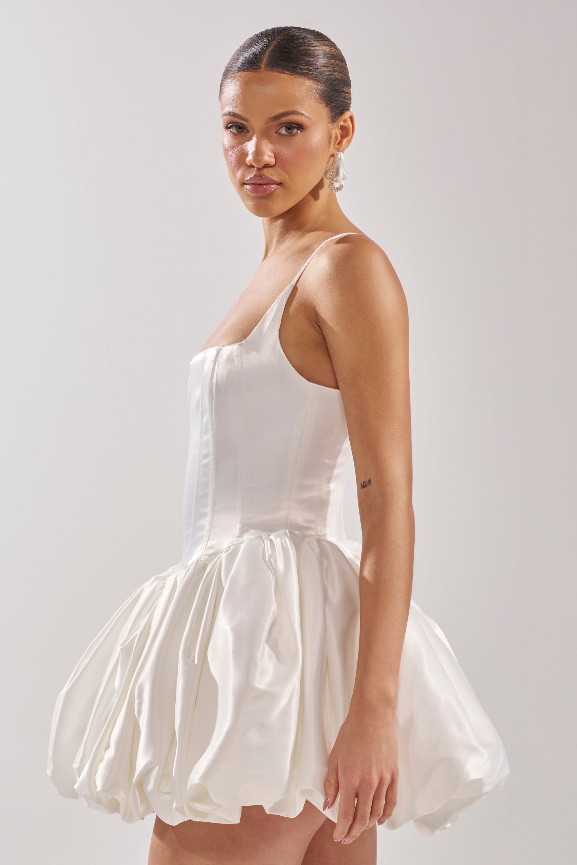 A woman with slicked-back hair models the JULIET SATIN MINI DRESS IN IVORY—featuring a corset bodice and voluminous ruffled bubble hem skirt—standing sideways and looking at the camera against a plain light background.