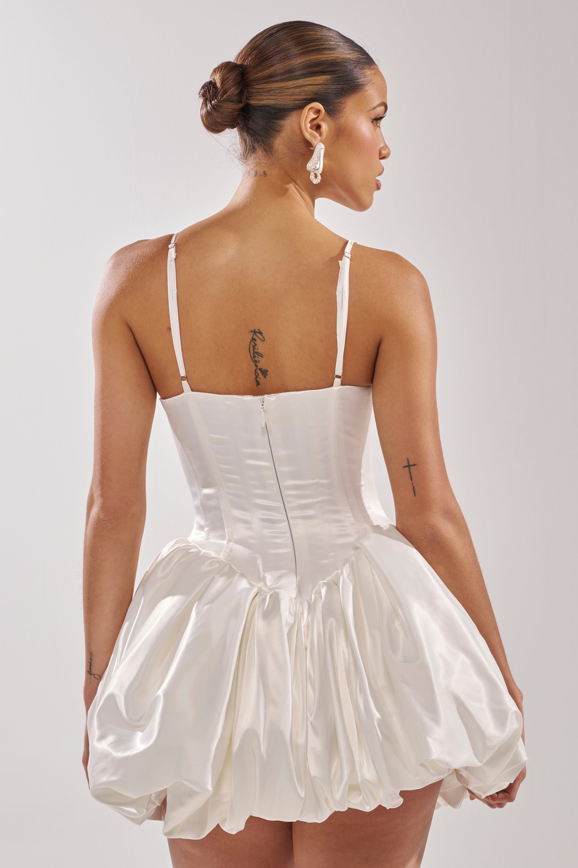 A woman with slicked-back hair models the JULIET SATIN MINI DRESS IN IVORY, featuring a corset bodice and bubble skirt. Facing away, she reveals tattoos on her upper back and right arm and wears large dangling earrings.