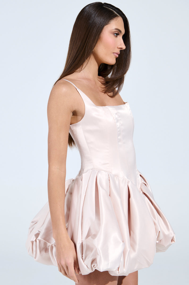 A woman with long brown hair poses in profile against a light background, wearing the Juliet Satin Mini Dress in Light Pink, showcasing its fitted bodice and flouncy bubble skirt.