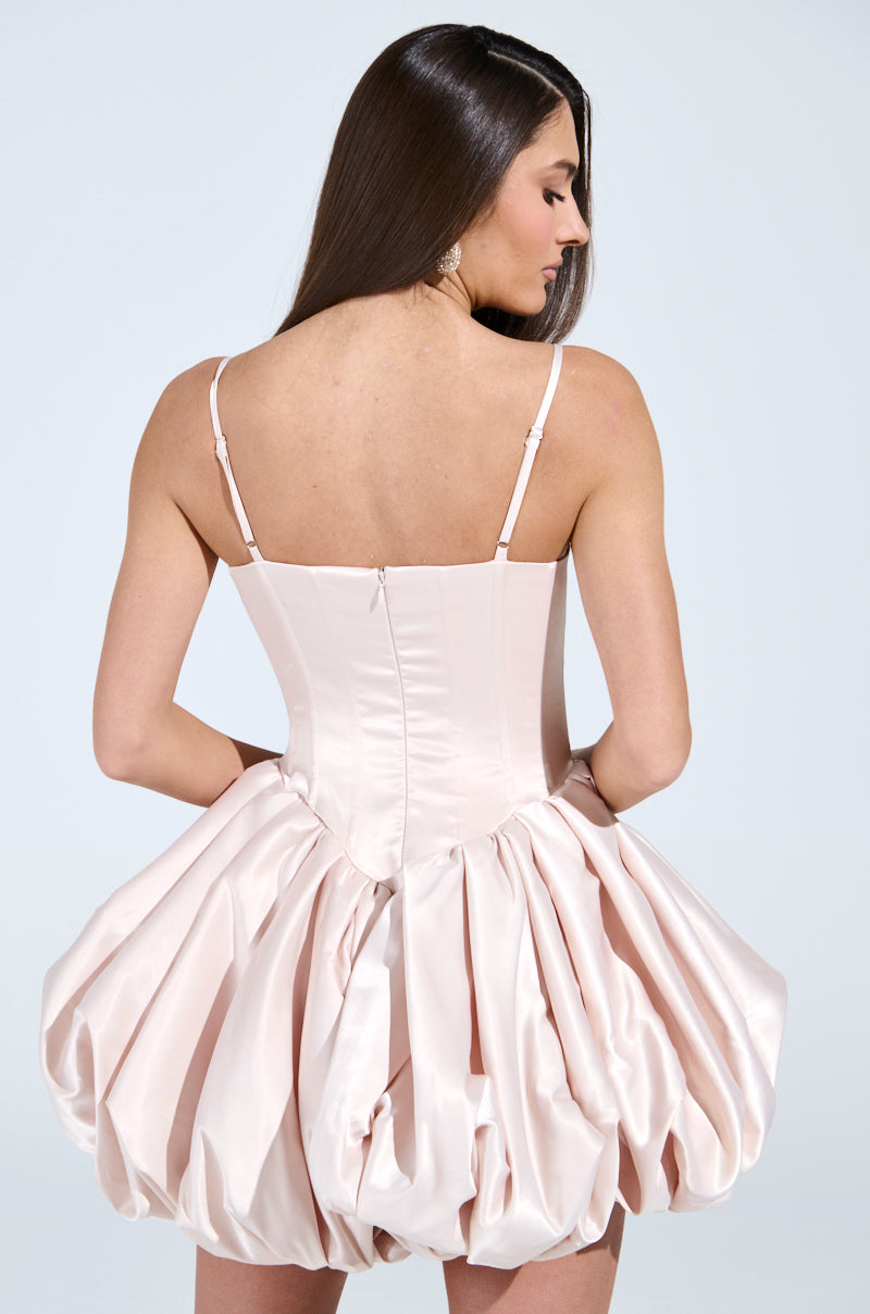 A woman with long hair wears the JULIET SATIN MINI DRESS IN LIGHT PINK, showcasing a structured bodice and flouncy bubble skirt. The dress fits snugly at the top and flares elegantly at the waist as she stands with her back to the camera.