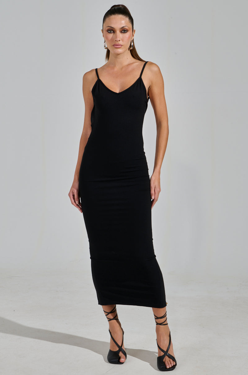 A woman poses against a plain backdrop in the MY GOODIES SEAMLESS MAXI DRESS, showcasing its bodycon silhouette. The fitted black dress has thin straps and a V-neckline, perfectly paired with black strappy heels. Her hair is elegantly pulled back to complete the look.