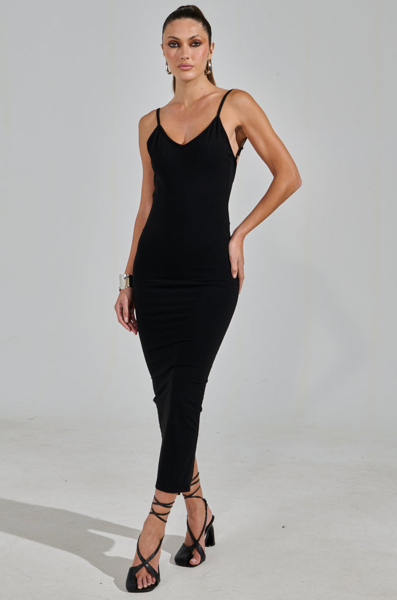 In a minimalist studio, a woman exudes confidence wearing the MY GOODIES SEAMLESS MAXI DRESS, a form-fitting bodycon with spaghetti straps. She completes her look with strappy black heels, a silver bracelet, and elegantly pulled-back hair.